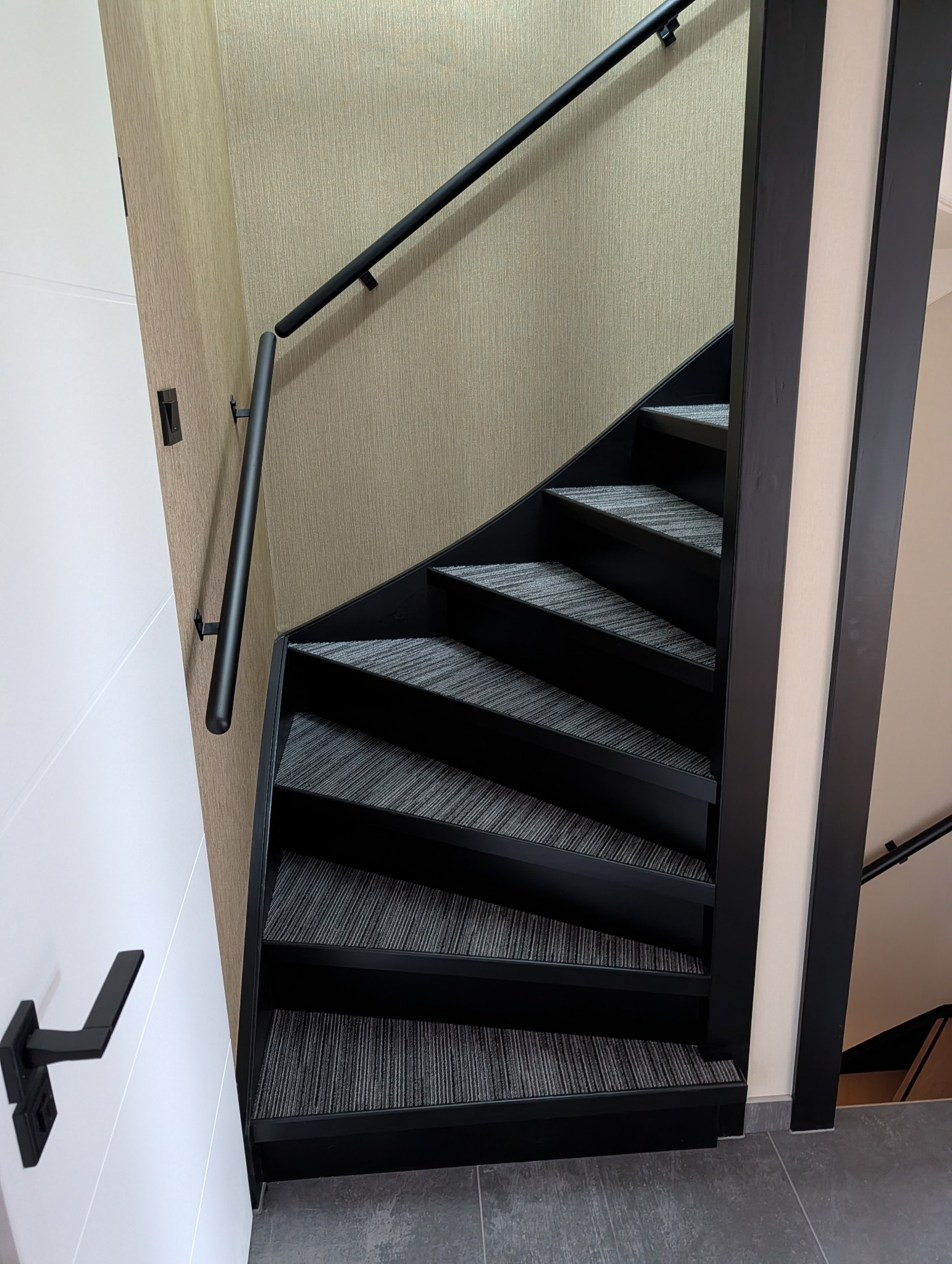 Nowoczesne, czarne schody z szarym dywanem na stopniach, widok z góry. Minimalistyczna balustrada i drzwi w tle. Wykończenie w neutralnych kolorach.