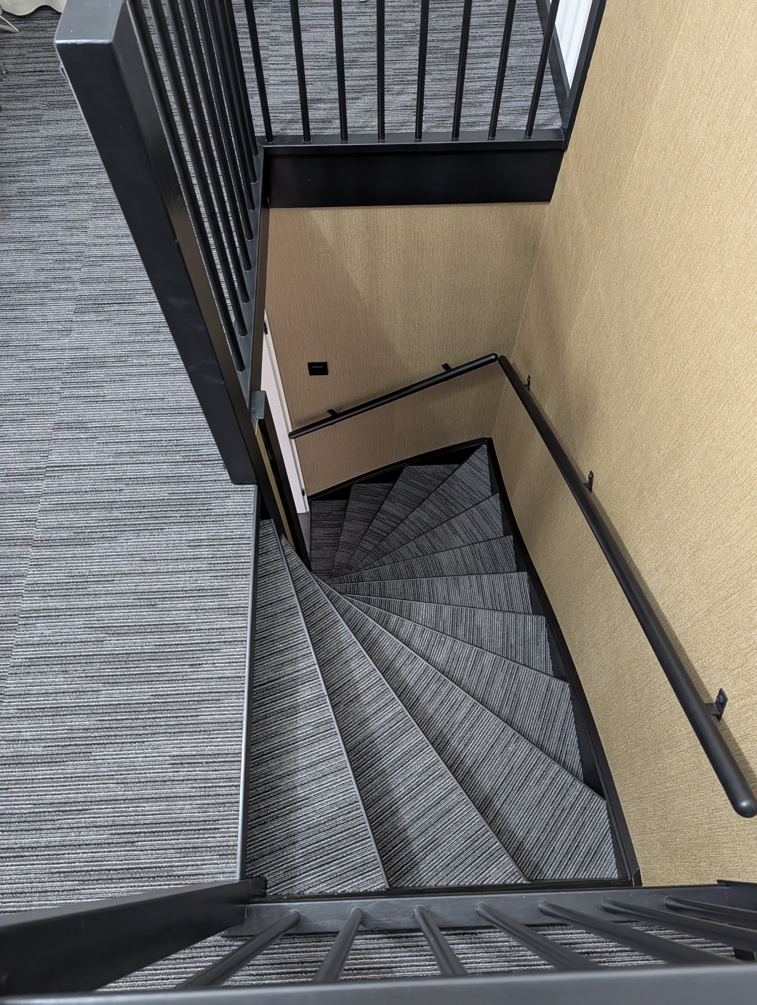 Spiralne schody z szarą wykładziną dywanową, widok z góry. Czarne metalowe balustrady i beżowa ściana. Nietypowy kąt ujęcia podkreśla geometrię schodów.