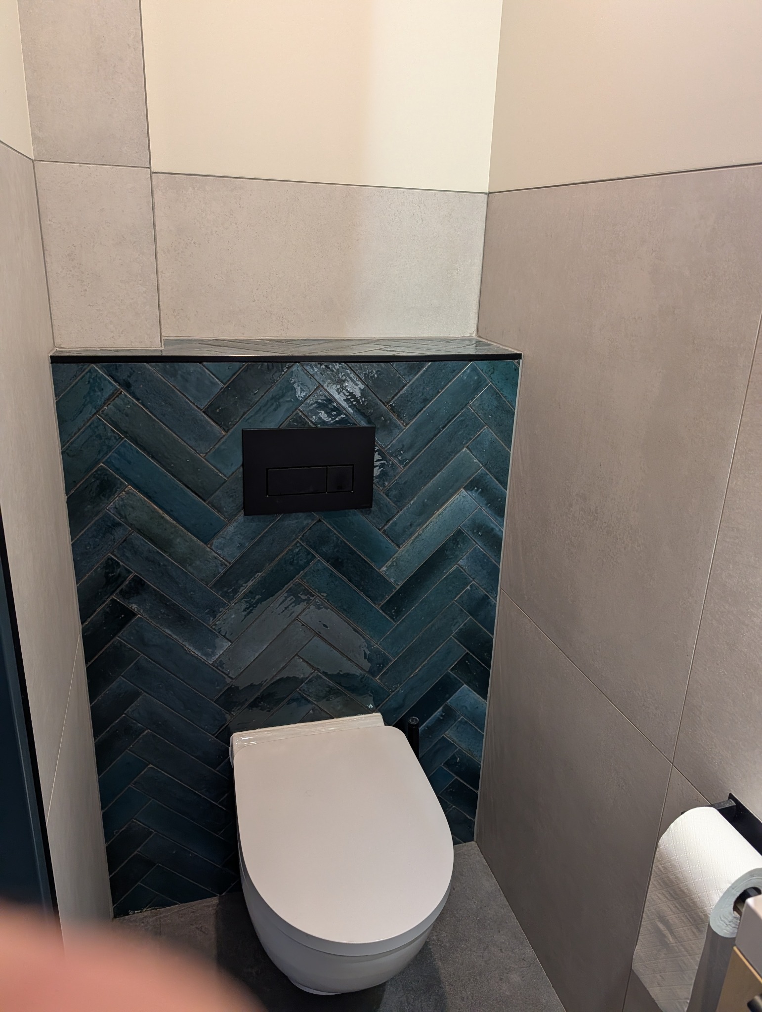 Nowoczesna toaleta podwieszana z niebieską mozaiką ułożoną w jodełkę i czarnym przyciskiem spłukującym, otoczona jasnymi płytkami. Minimalistyczny design.