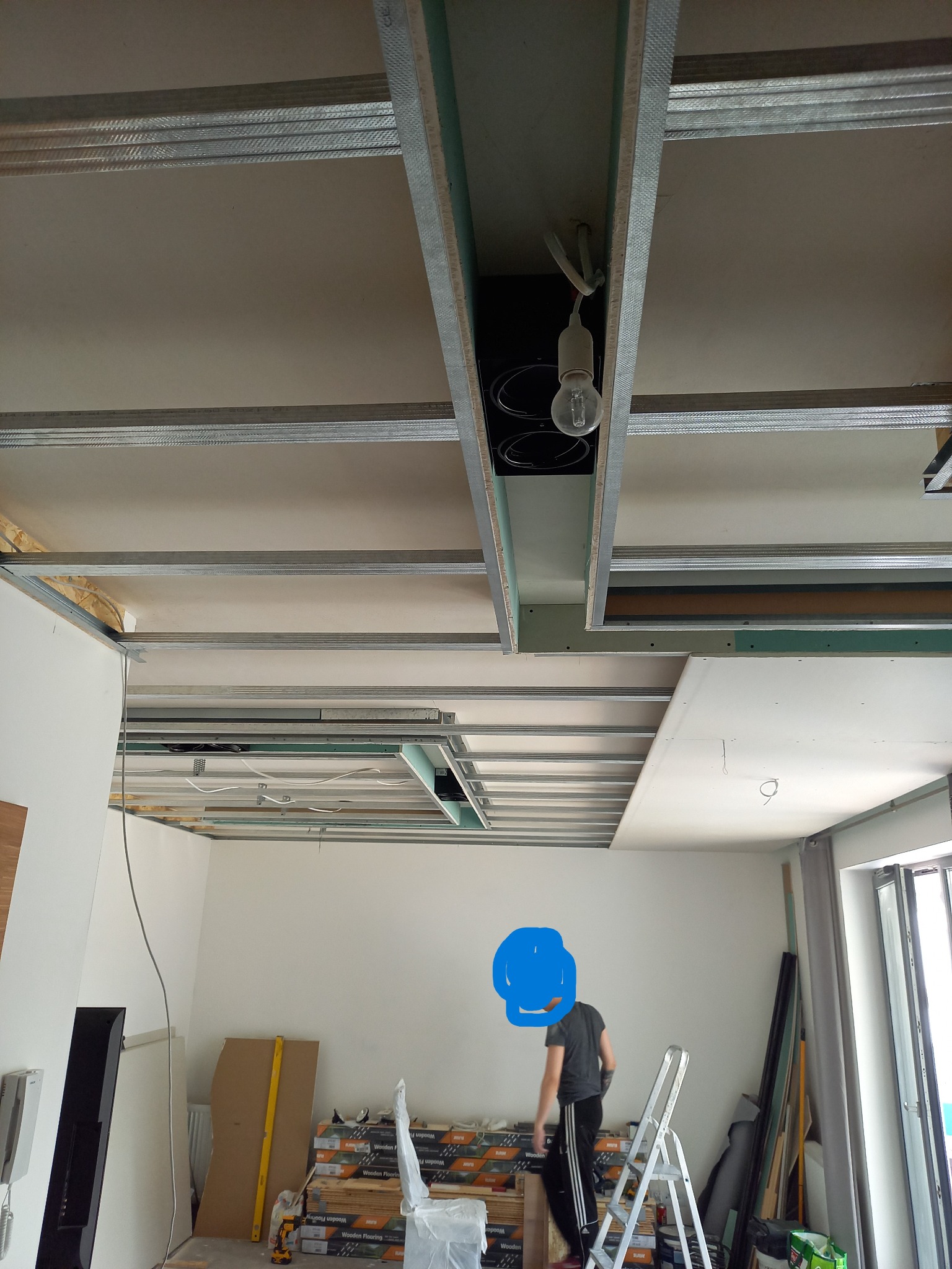 Montaż sufitu podwieszanego z płyt kartonowo-gipsowych. Widoczne metalowe profile, częściowo zamontowane płyty i instalacja oświetleniowa. W tle drabina i materiały budowlane.