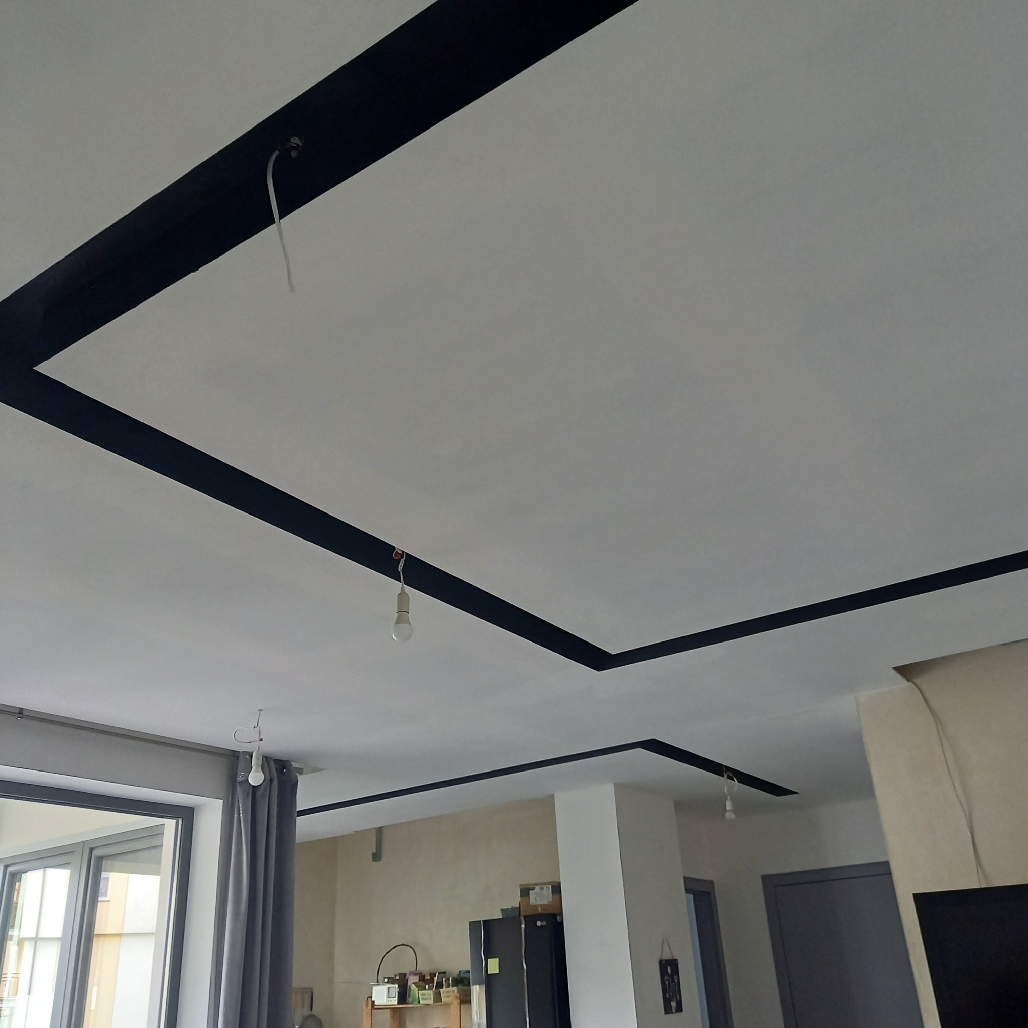 Biały sufit z czarnymi, geometrycznymi pasami dekoracyjnymi, widoczne żarówki na kablach, fragment okna i wyposażenia wnętrza. Minimalistyczny design.