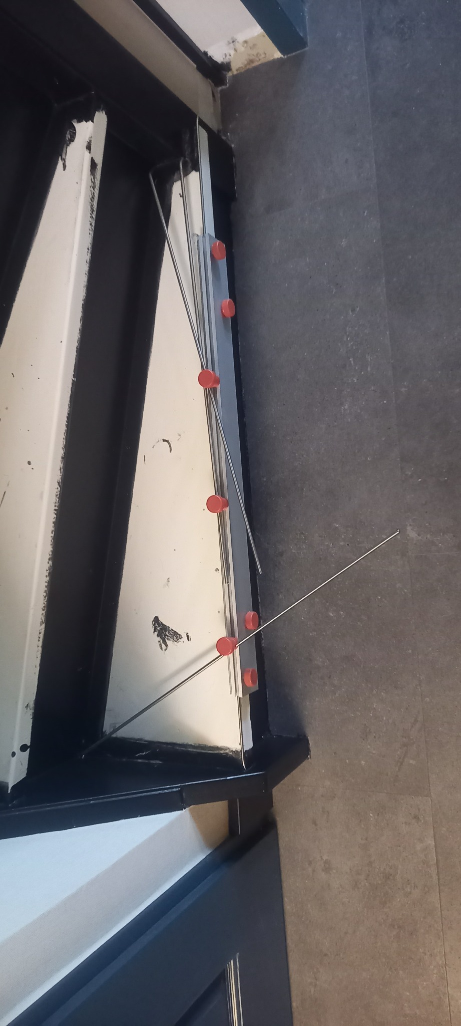 Fragment schodów z zamontowanymi listwami i czerwonymi podkładkami dystansowymi, przygotowanymi do montażu płytek. Widok z góry na szare płytki podłogowe.