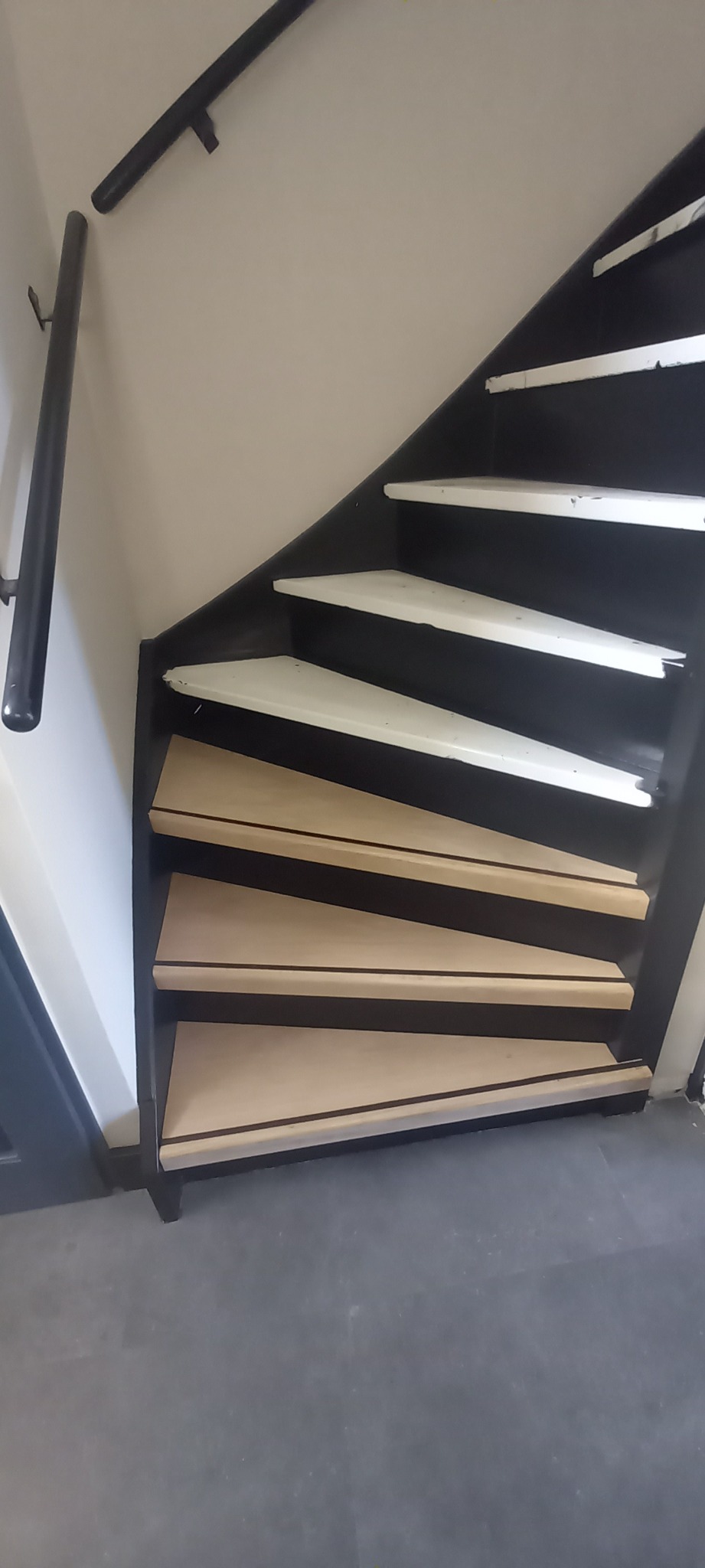 Spiralne schody z czarną ramą i drewnianymi stopniami, częściowo pomalowanymi na biało, widziane z dołu, z fragmentem poręczy na ścianie.