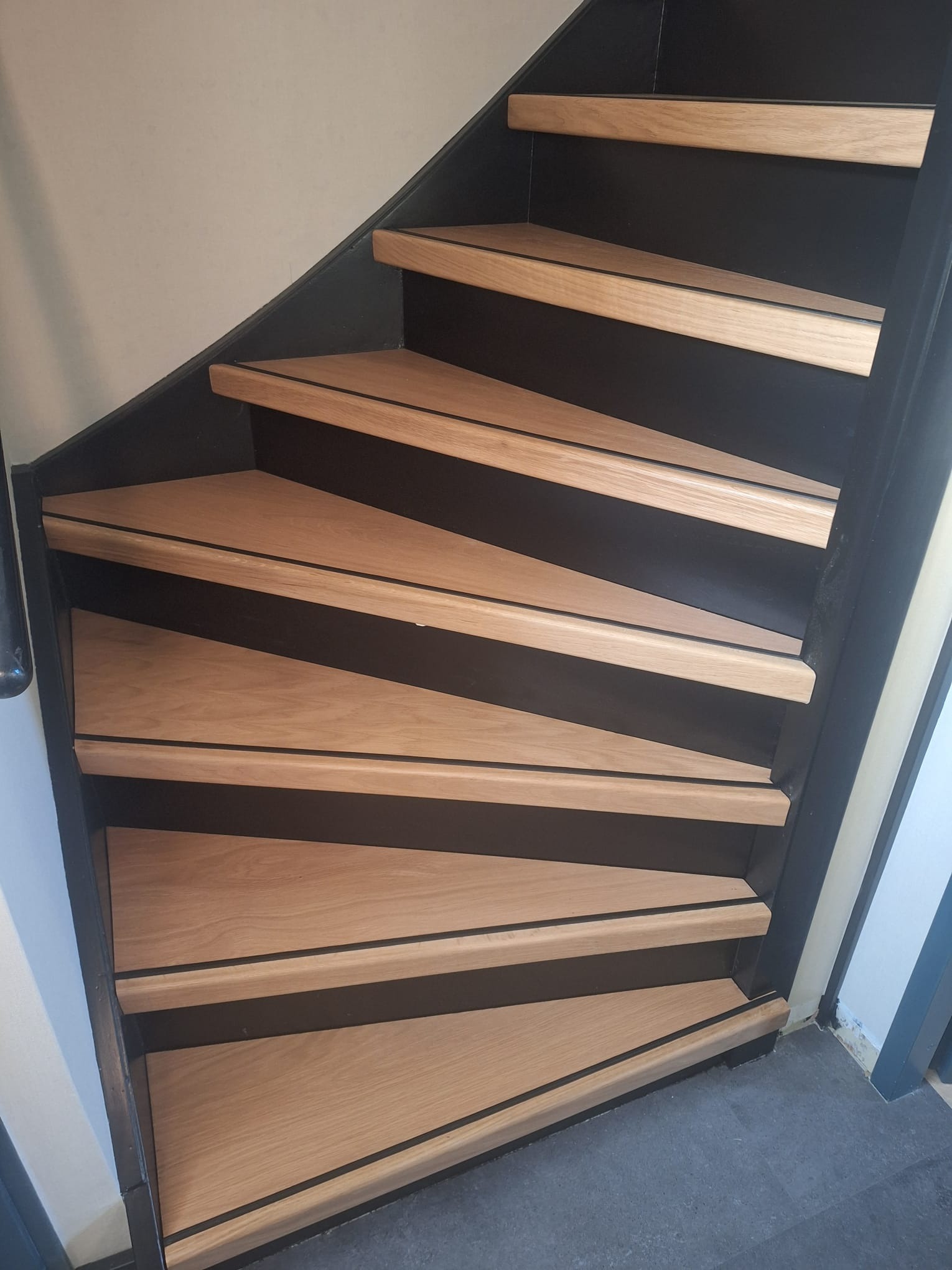 Drewniane schody z czarnymi elementami konstrukcyjnymi, widok z dołu, ukazujący detale wykończenia i kąt ułożenia stopni.