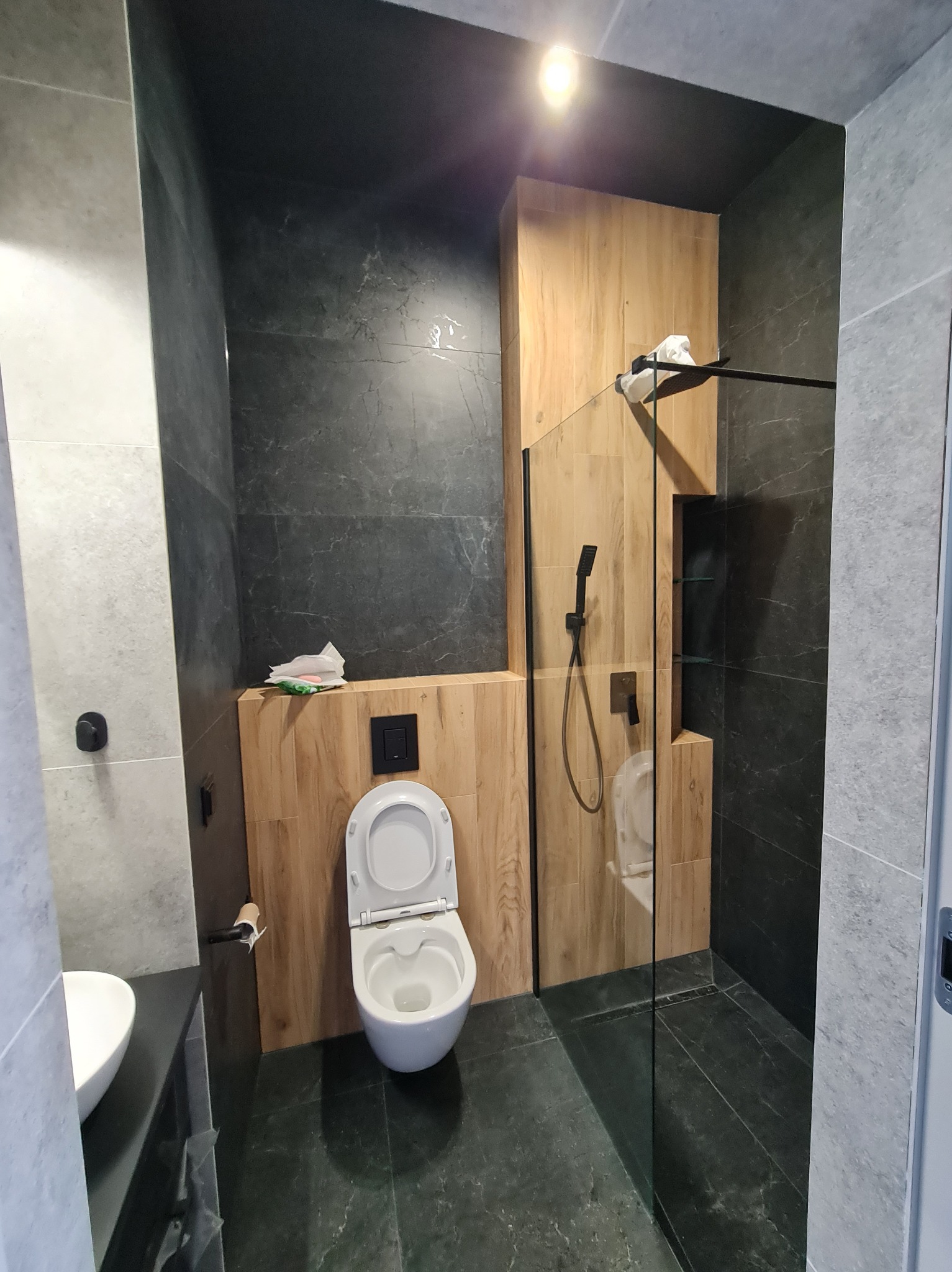 Nowoczesna łazienka z drewnianymi akcentami: kabina prysznicowa ze szklaną ścianką, podwieszana toaleta i umywalka na tle ciemnych, marmurkowych płytek.