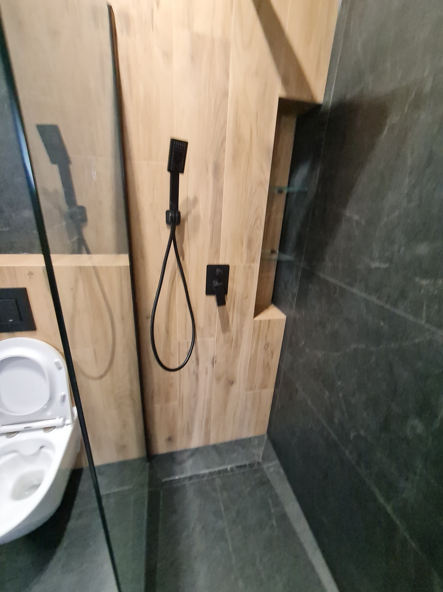 Nowoczesna łazienka z kabiną typu walk-in, wykończona płytkami imitującymi drewno i szarym kamieniem. Widoczna toaleta i czarna armatura.