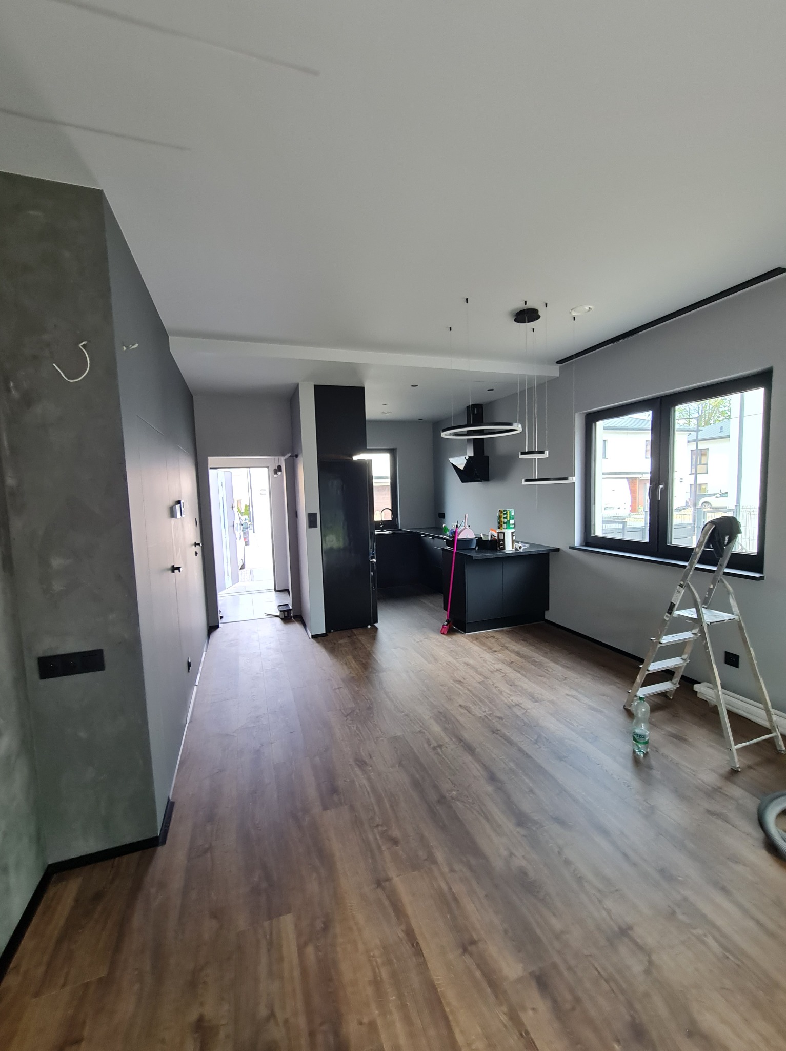 Nowoczesne wnętrze po remoncie: szare ściany z efektem betonu, drewniana podłoga, czarna kuchnia. Widoczna drabina i butelka wody.