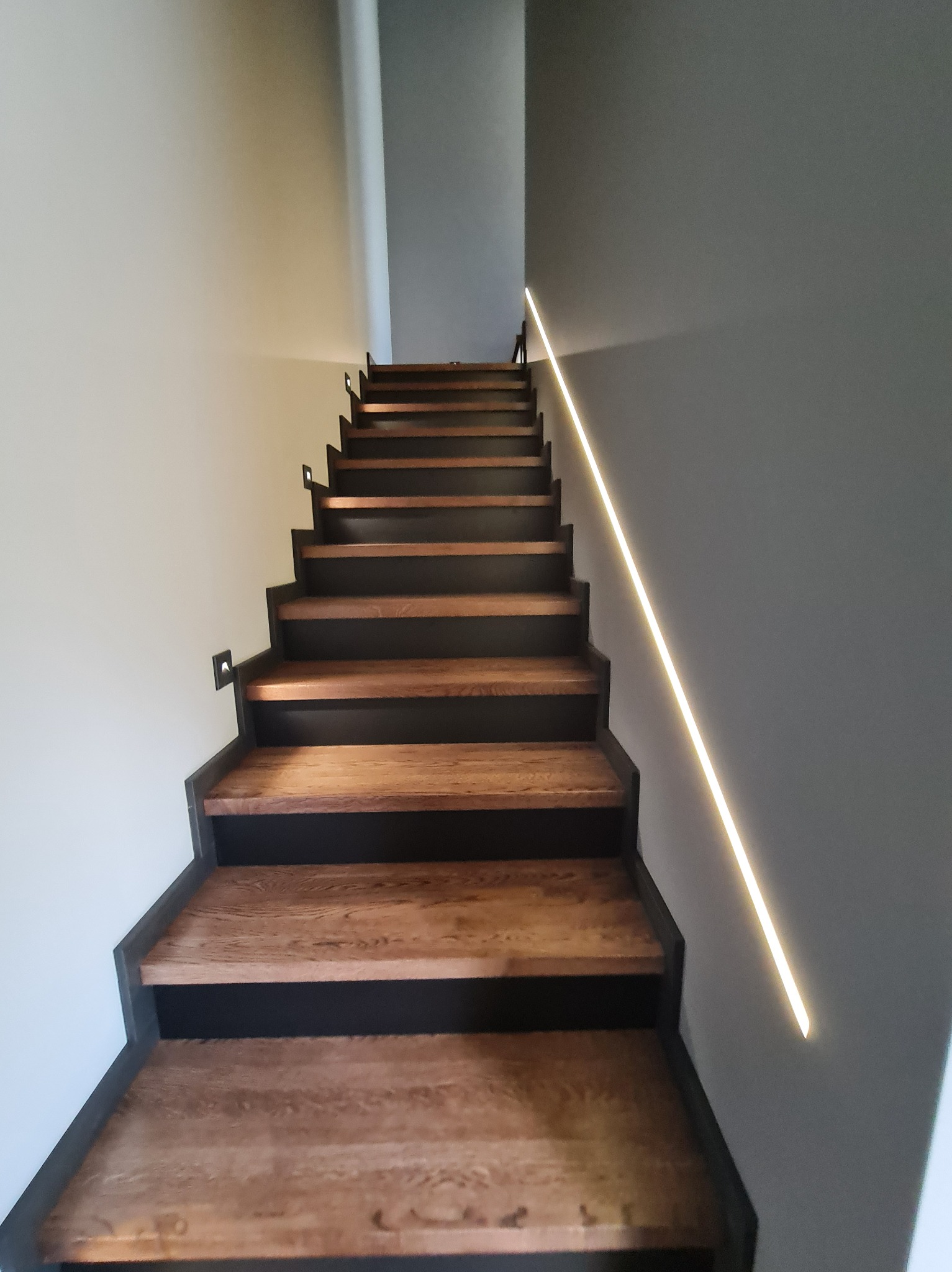 Nowoczesne schody drewniane z czarnymi podstopnicami i subtelnym oświetleniem LED wzdłuż ściany, widok z dołu, minimalistyczny design.
