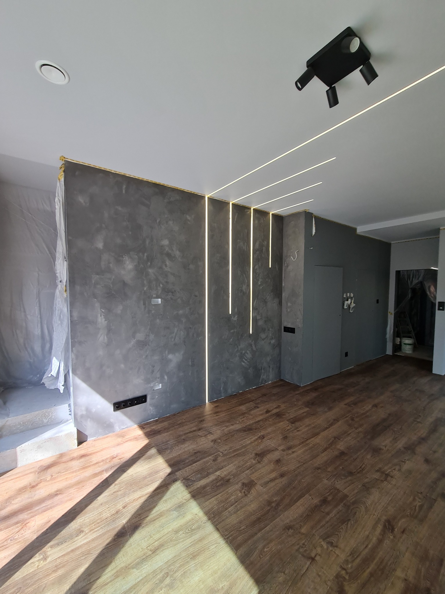 Wykończone wnętrze pokoju z szarą ścianą o fakturze betonu, oświetleniem LED i drewnianą podłogą. Część pomieszczenia w trakcie prac wykończeniowych.