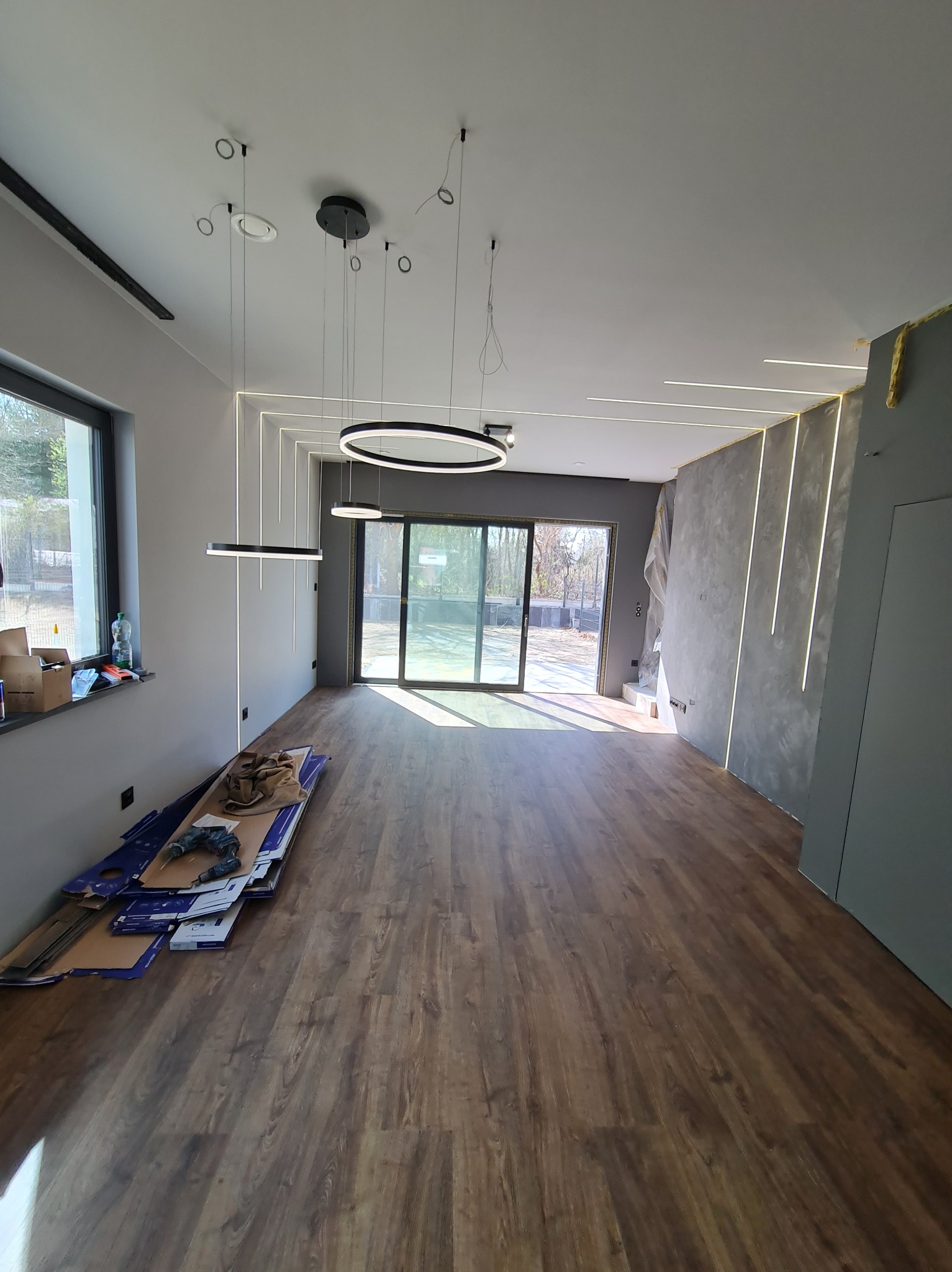 Nowoczesne wnętrze po remoncie: drewniana podłoga, szare ściany z oświetleniem LED, okrągłe lampy wiszące. Widoczne narzędzia i materiały budowlane.