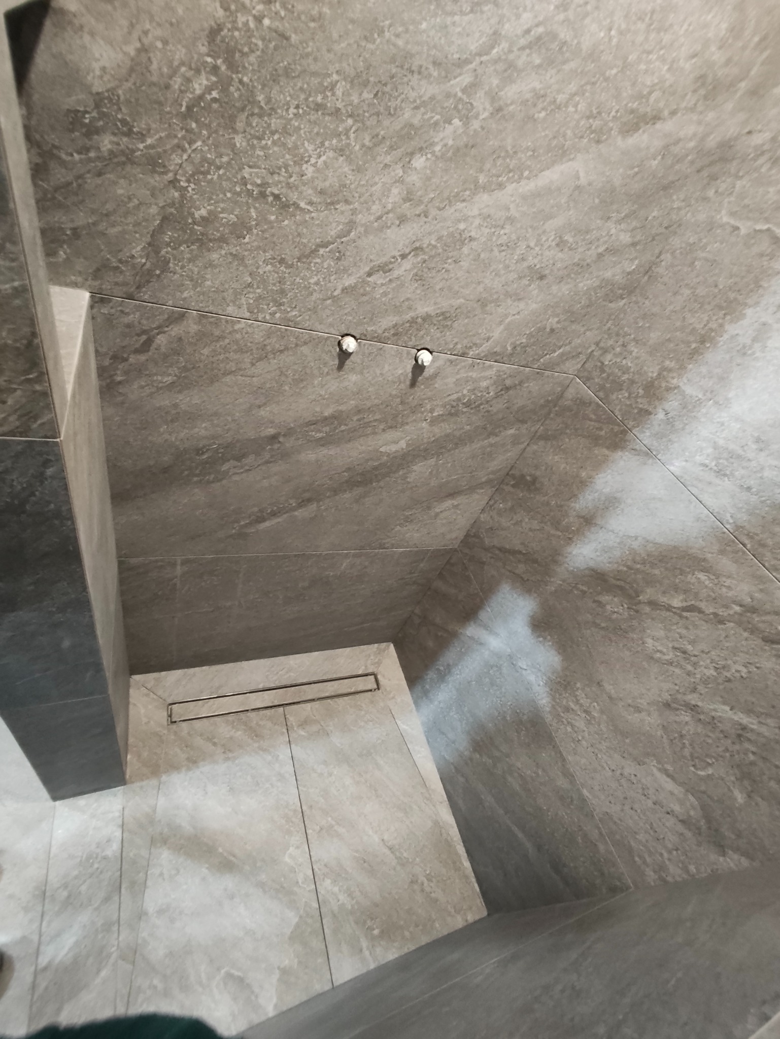 Wykończona łazienka z szarymi płytkami imitującymi kamień, widoczny odpływ liniowy i dwa punkty mocowania na ścianie. Minimalistyczny design.