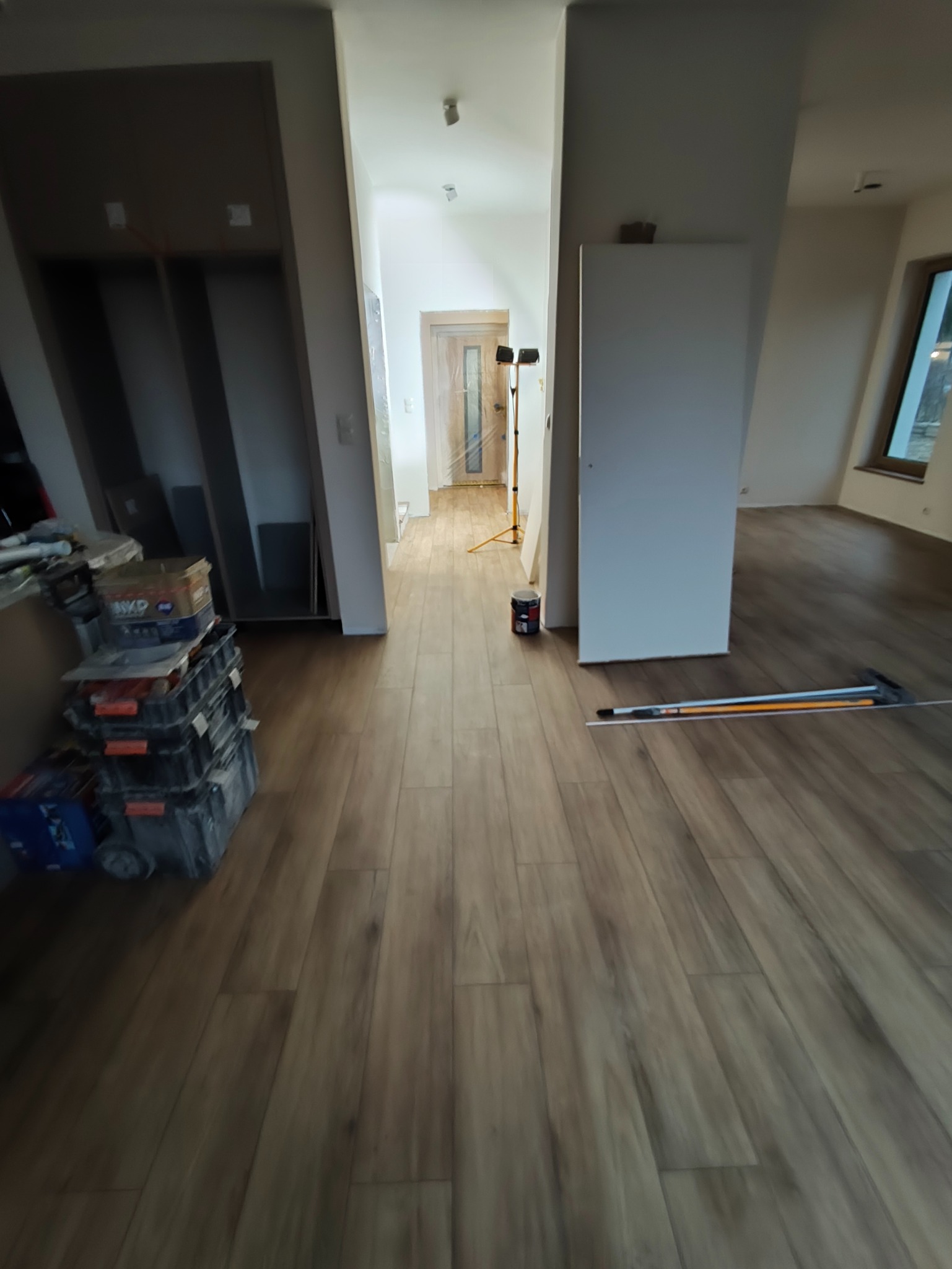 Ułożona podłoga z paneli w odcieniach brązu, widok na korytarz z otwartymi drzwiami, narzędzia pracy i pojemniki na podłodze, wnętrze w trakcie wykańczania.