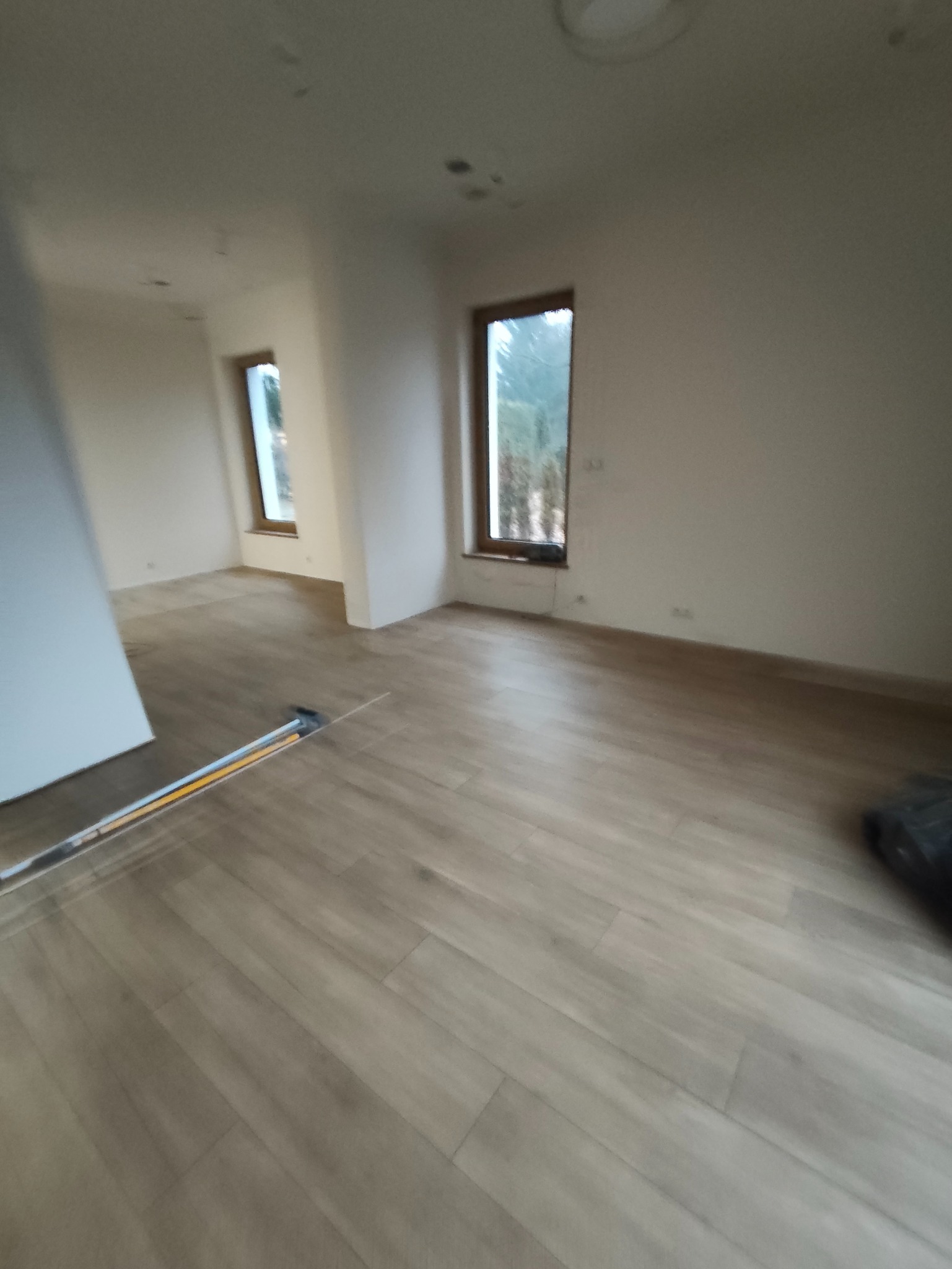 Jasne panele podłogowe w nowym wnętrzu z dwoma oknami. Ściany w odcieniach bieli, widoczne listwy przypodłogowe. Minimalistyczny design, świeżo po remoncie.