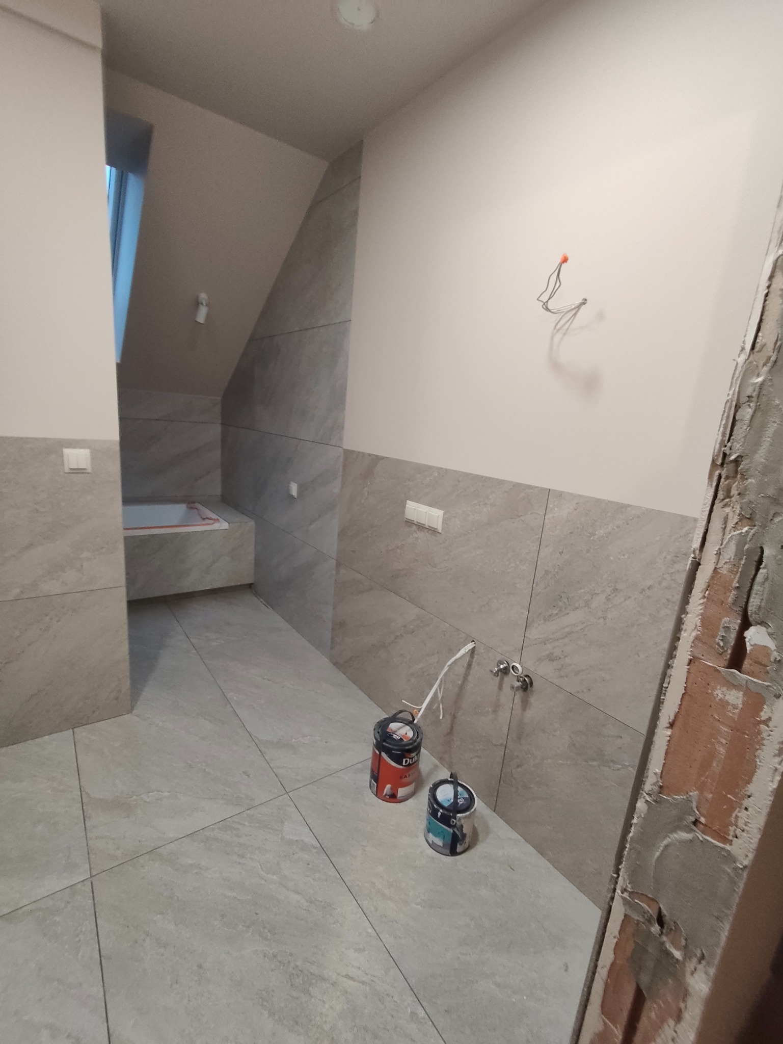 Wykończona łazienka z szarymi płytkami, widoczne puszki z farbą i fragment ściany w trakcie prac. Ujęcie z perspektywy podłogi, podkreślające detale wykończenia.