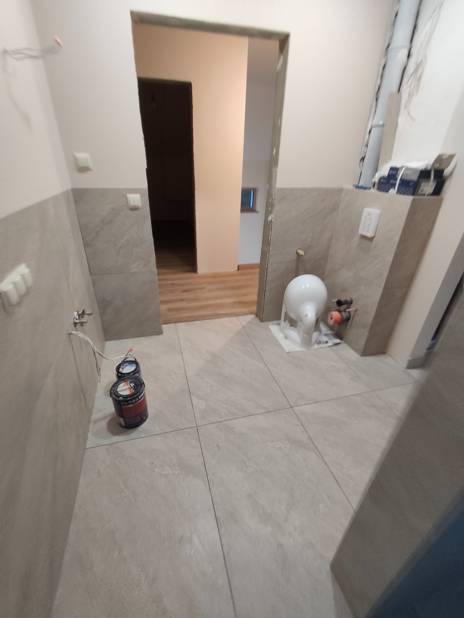 Wykończona łazienka z beżowymi płytkami, widoczne puszki z farbą i częściowo zamontowana toaleta. W tle widoczne drewniane panele podłogowe w innym pomieszczeniu.