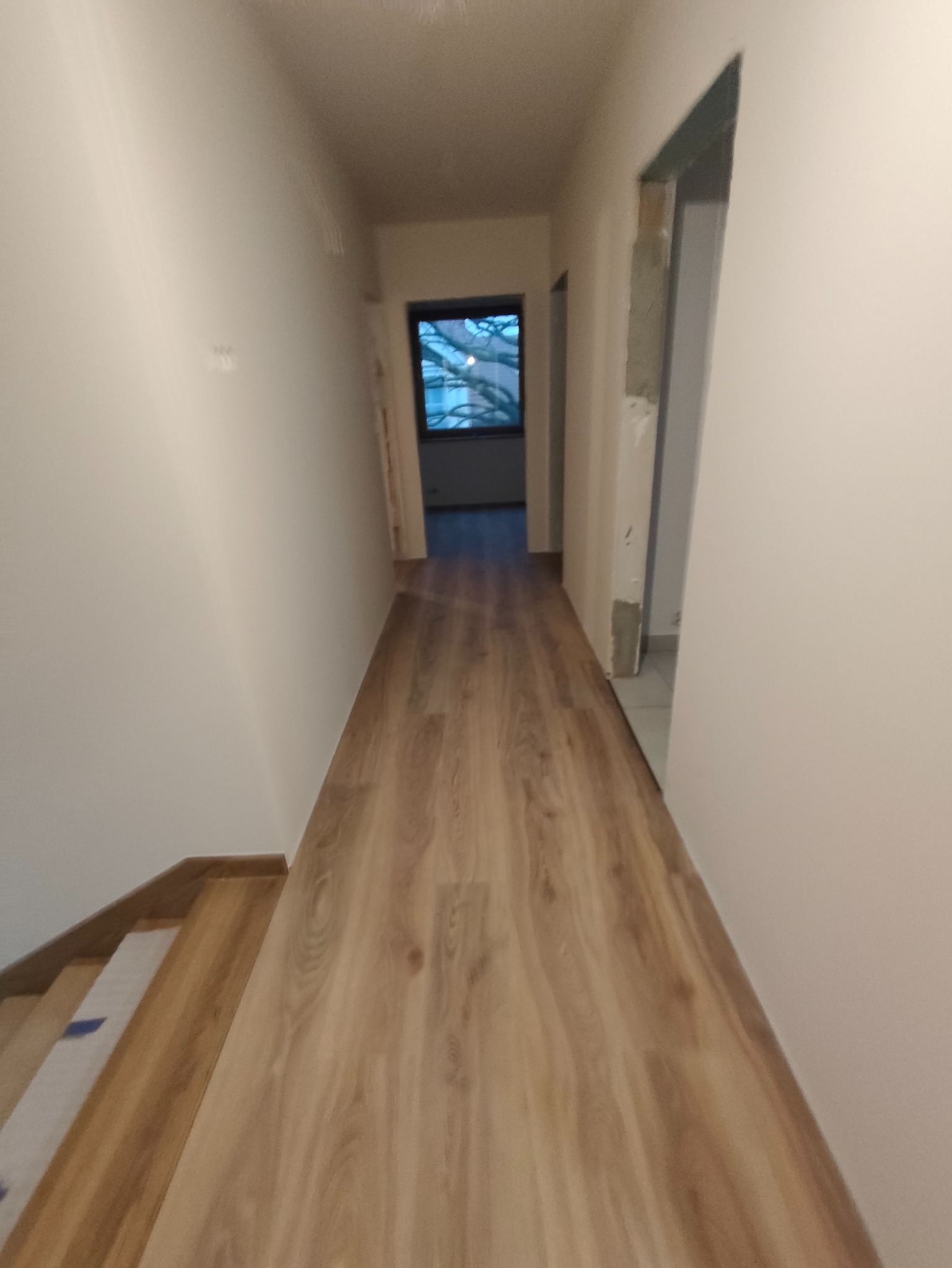 Korytarz po remoncie z nową podłogą imitującą drewno. Widoczne drzwi w trakcie montażu i okno na końcu korytarza. Ściany pomalowane na jasny kolor.