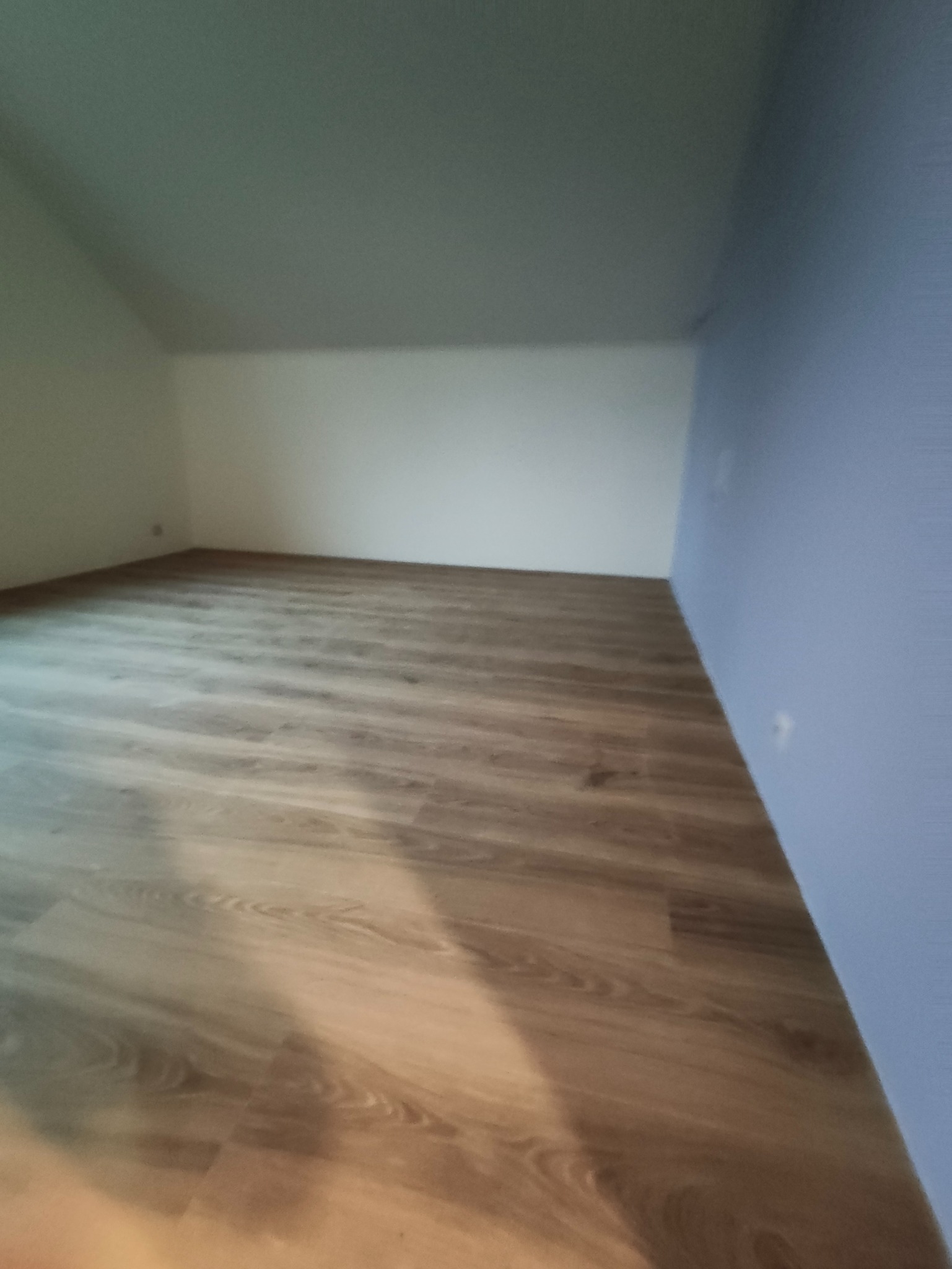 Puste pomieszczenie po remoncie: świeżo położona podłoga z paneli imitujących drewno, białe i niebieskie ściany, skośny sufit. Widok na całe pomieszczenie.