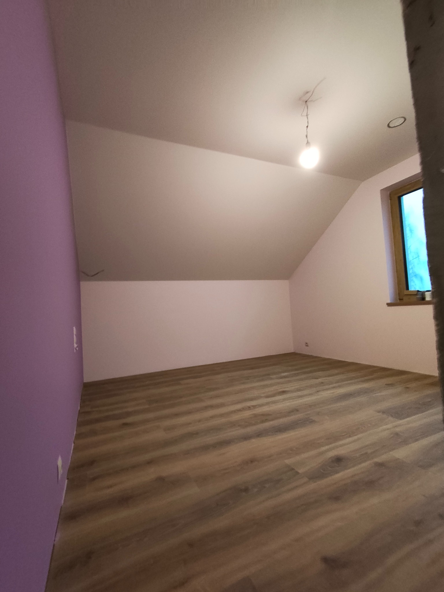 Poddasze po remoncie: świeżo pomalowane ściany w odcieniach fioletu i beżu, drewniana podłoga, okno. Widoczna surowa żarówka. Ukończone wykończenie wnętrza.