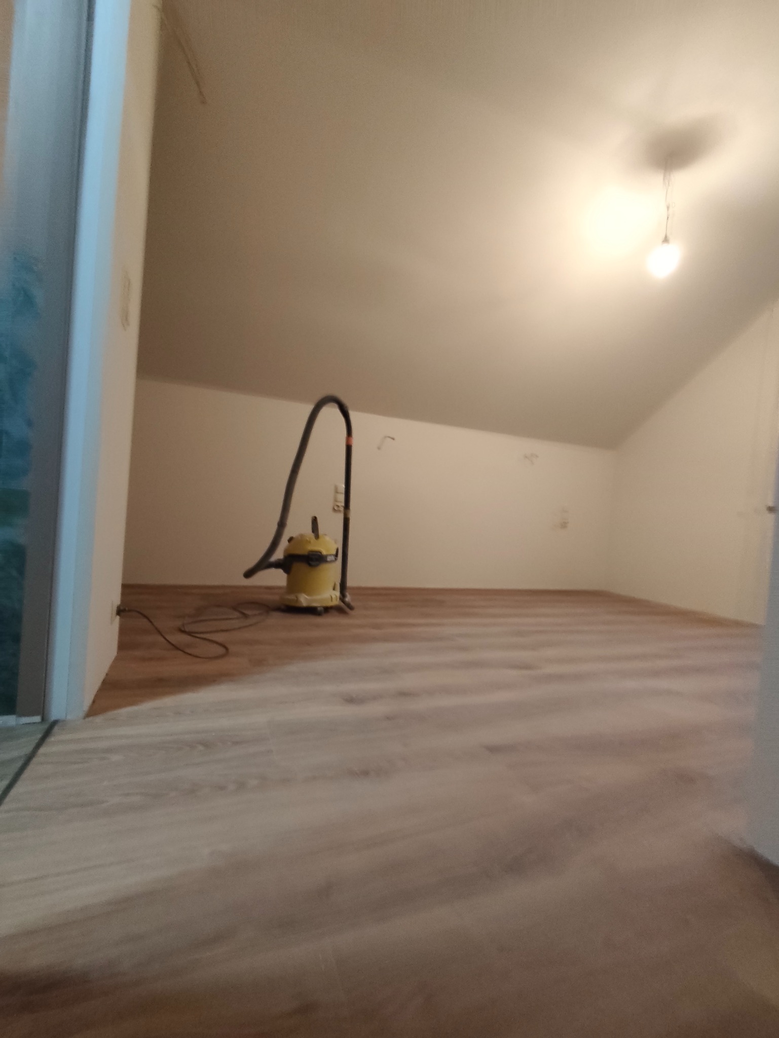 Poddasze po remoncie z nową podłogą, widoczny odkurzacz przemysłowy. Świeżo wykończone wnętrze z jasnymi ścianami i podłogą w odcieniach drewna.