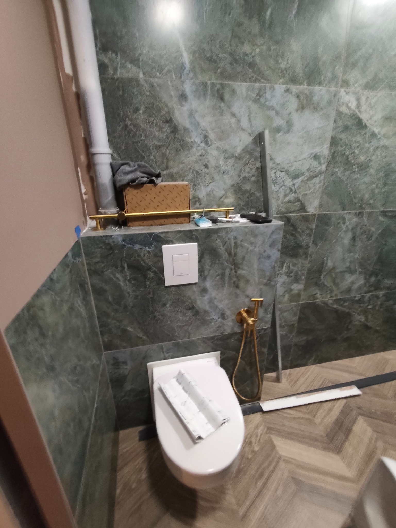 Wykończona łazienka z marmurowymi płytkami, złotą armaturą i toaletą z deską. Na podłodze parkiet ułożony w jodełkę. Widoczny fragment instalacji.