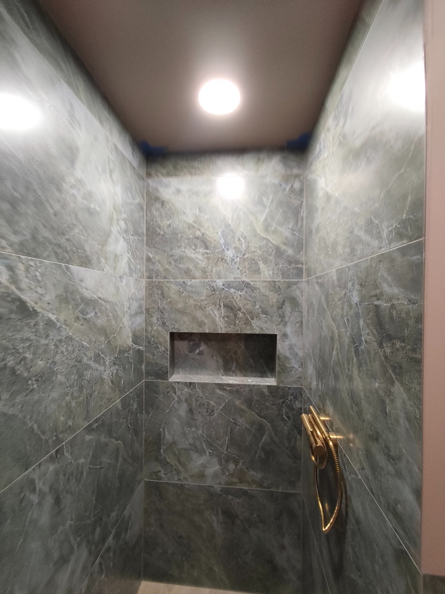 Wykończona kabina prysznicowa z szarymi płytkami imitującymi marmur, wnęką w ścianie i złotą armaturą. Górne oświetlenie punktowe.