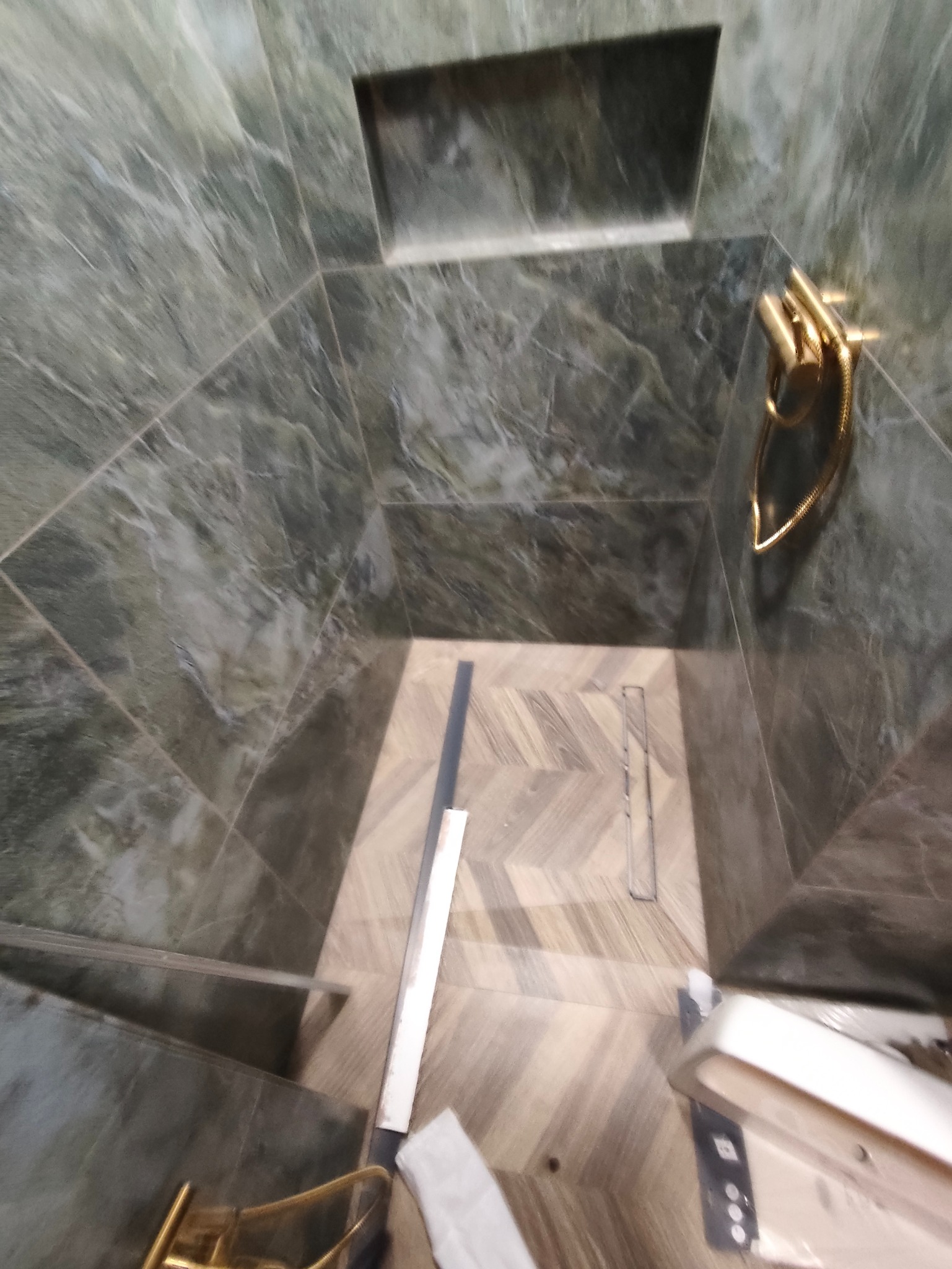 Wykończona kabina prysznicowa z zielonymi płytkami imitującymi marmur i podłogą w jodełkę. Widoczny odpływ liniowy i złota bateria.