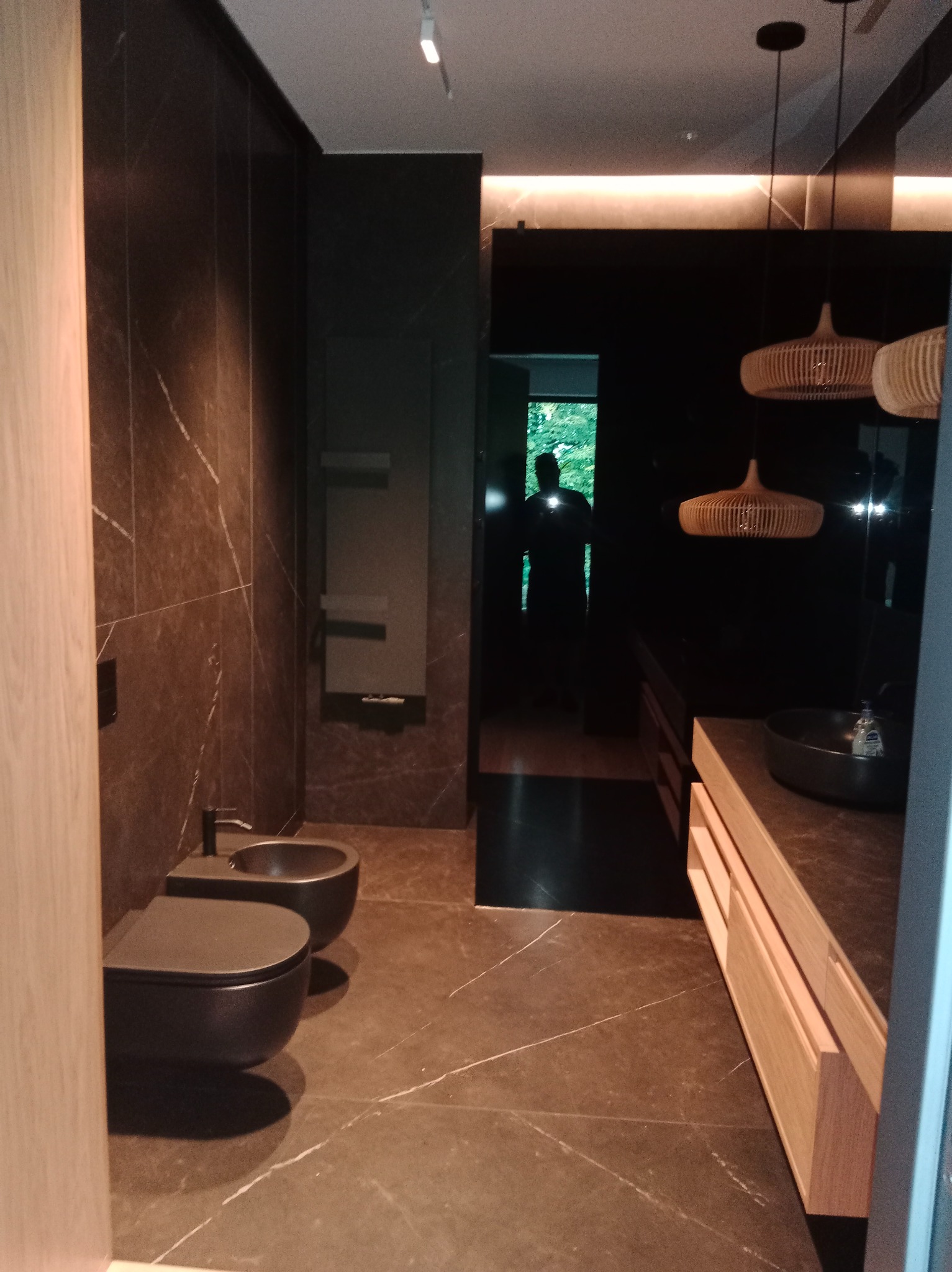 Nowoczesna łazienka z ciemnymi płytkami imitującymi marmur, podwieszaną toaletą i bidetem. Umywalka nablatowa i drewniane szafki podświetlone od góry.