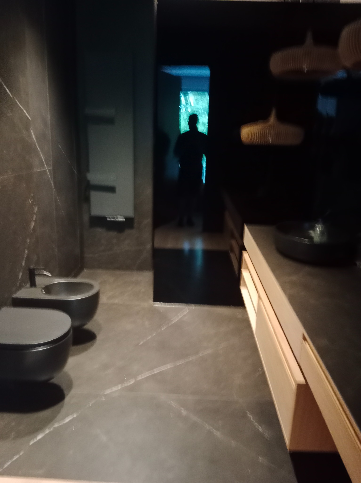 Nowoczesna łazienka z czarnymi płytkami imitującymi marmur, wiszącą toaletą i bidetem. Umywalka nablatowa na szafce z jasnego drewna. W tle sylwetka osoby w drzwiach.