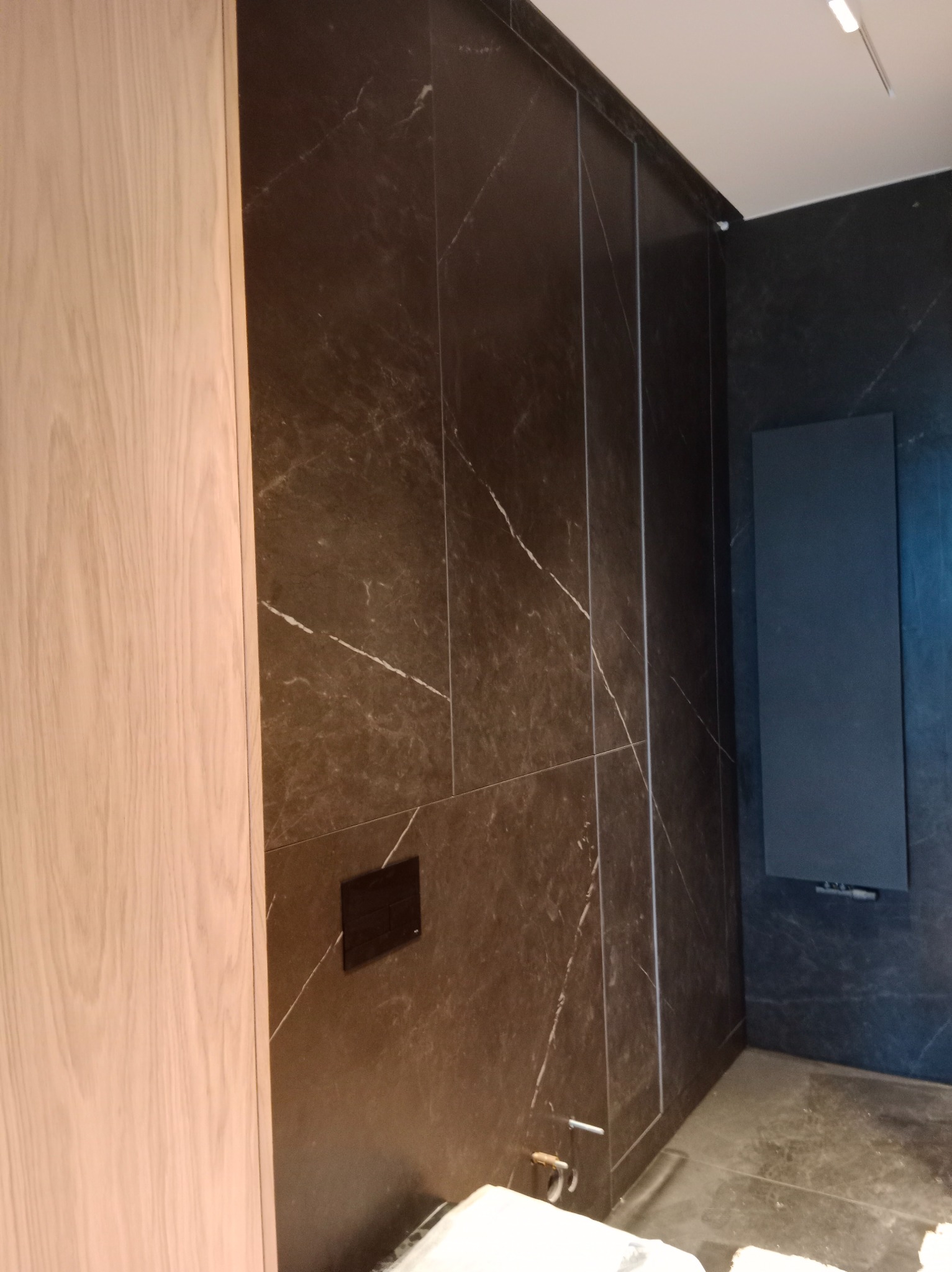 Elegancka łazienka z ciemnymi, marmurkowymi płytkami i drewnianą szafką. Widoczny grzejnik panelowy i czarny panel sterujący. Minimalistyczny design.