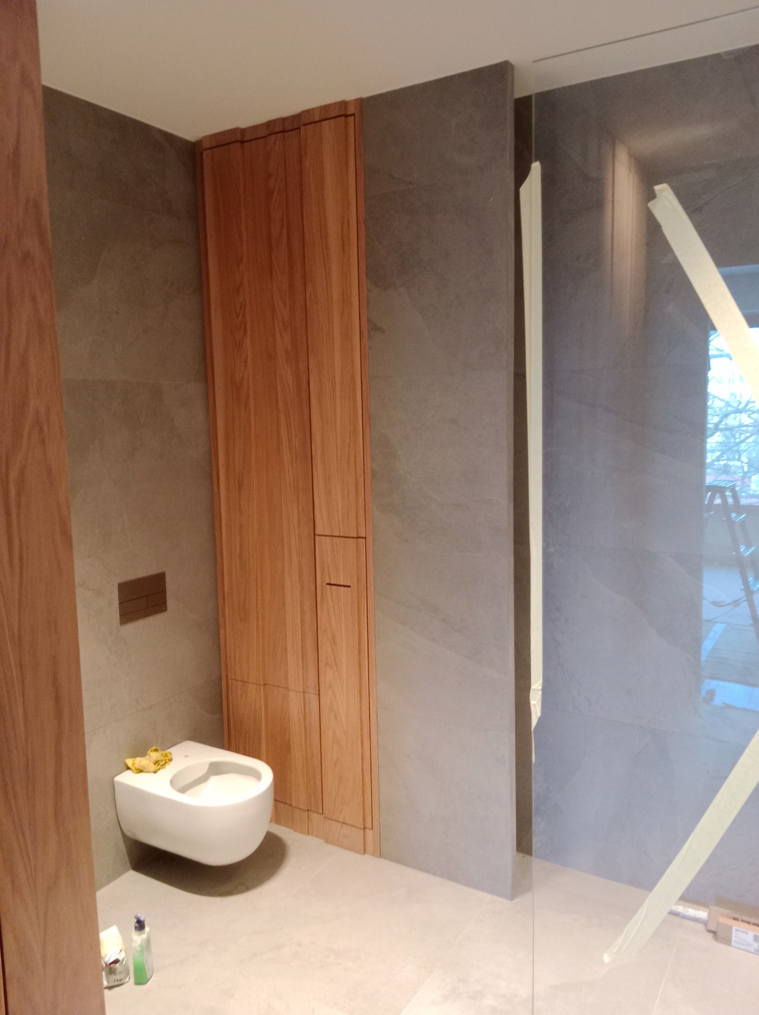 Nowoczesna łazienka z wiszącą toaletą, drewnianą zabudową i szarą płytką imitującą kamień. Widoczne akcesoria i zabezpieczenia na szklanej kabinie.