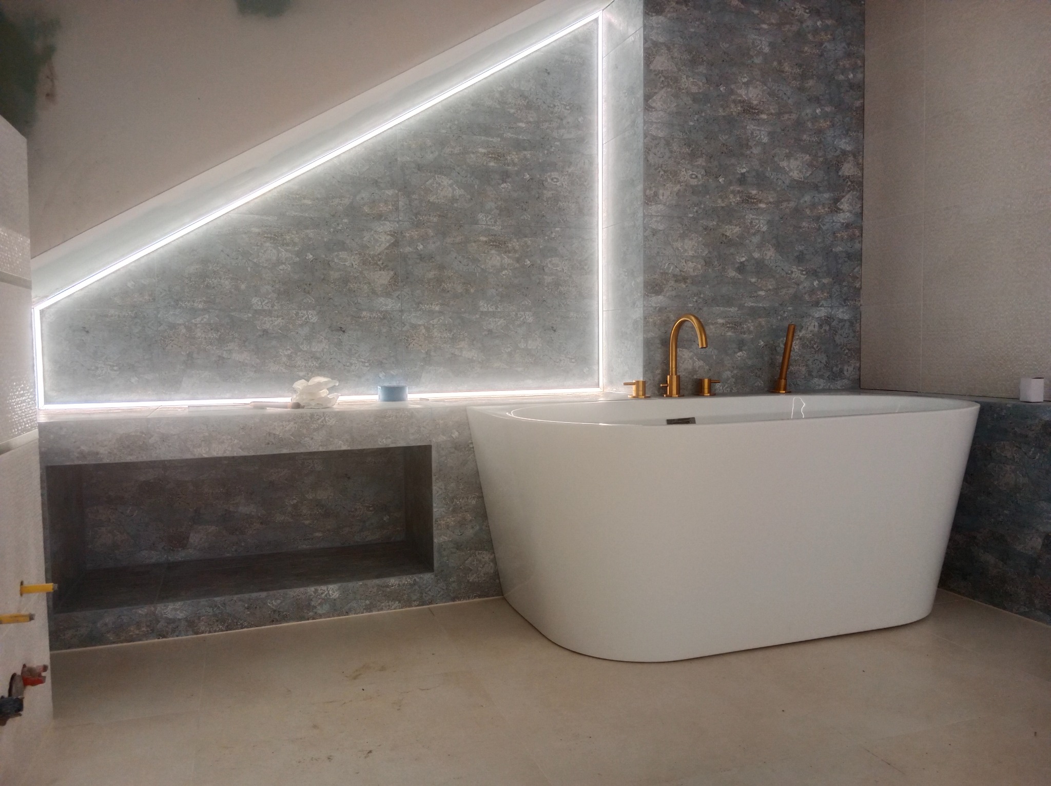 Nowoczesna łazienka z wolnostojącą białą wanną, złotą armaturą i podświetlaną wnęką w szaro-niebieskiej płytce. Minimalistyczny design, jasne płytki podłogowe.