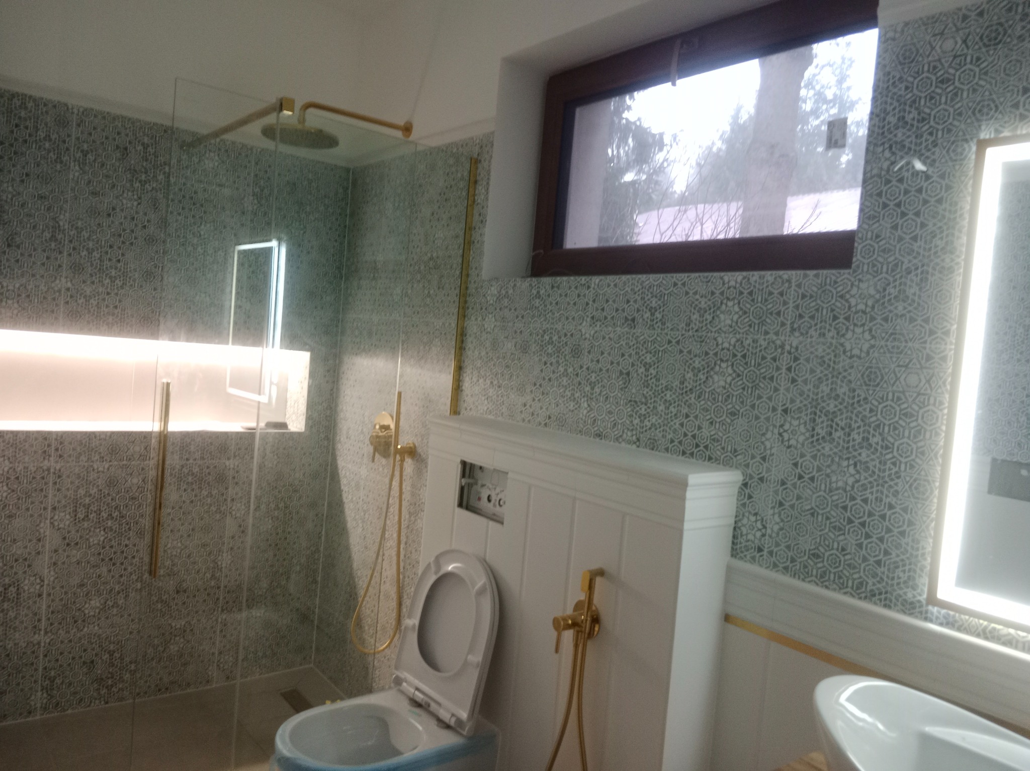 Wykończona łazienka z kabiną prysznicową typu walk-in, toaletą z zabudowaną spłuczką i umywalką. Płytki w geometryczny wzór, złote detale armatury i oświetlenia.