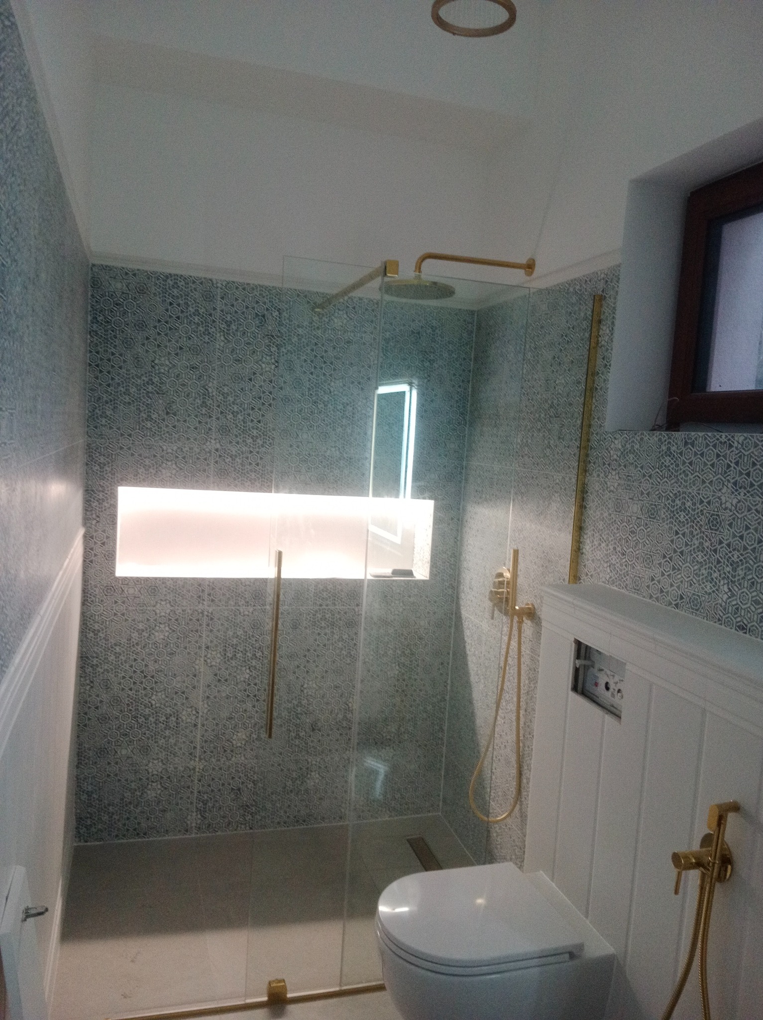 Nowoczesna łazienka z kabiną prysznicową typu walk-in, wykończona płytkami w geometryczny wzór i złotymi akcentami. Widoczna toaleta podwieszana i oświetlona wnęka.