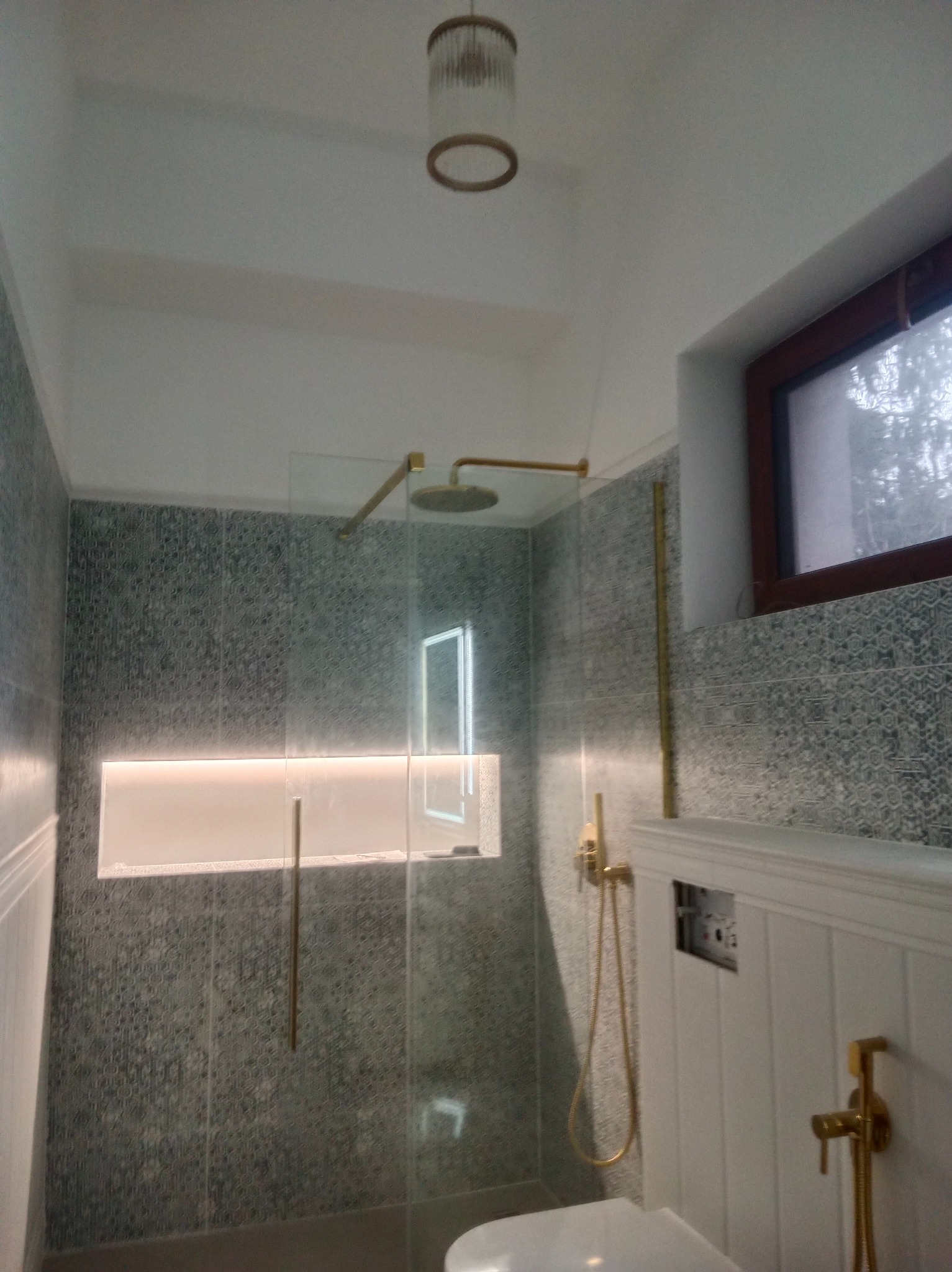 Nowoczesna łazienka z kabiną prysznicową typu walk-in, wykończona szaro-niebieską mozaiką i złotymi detalami armatury. Widoczne oświetlenie LED i okno.