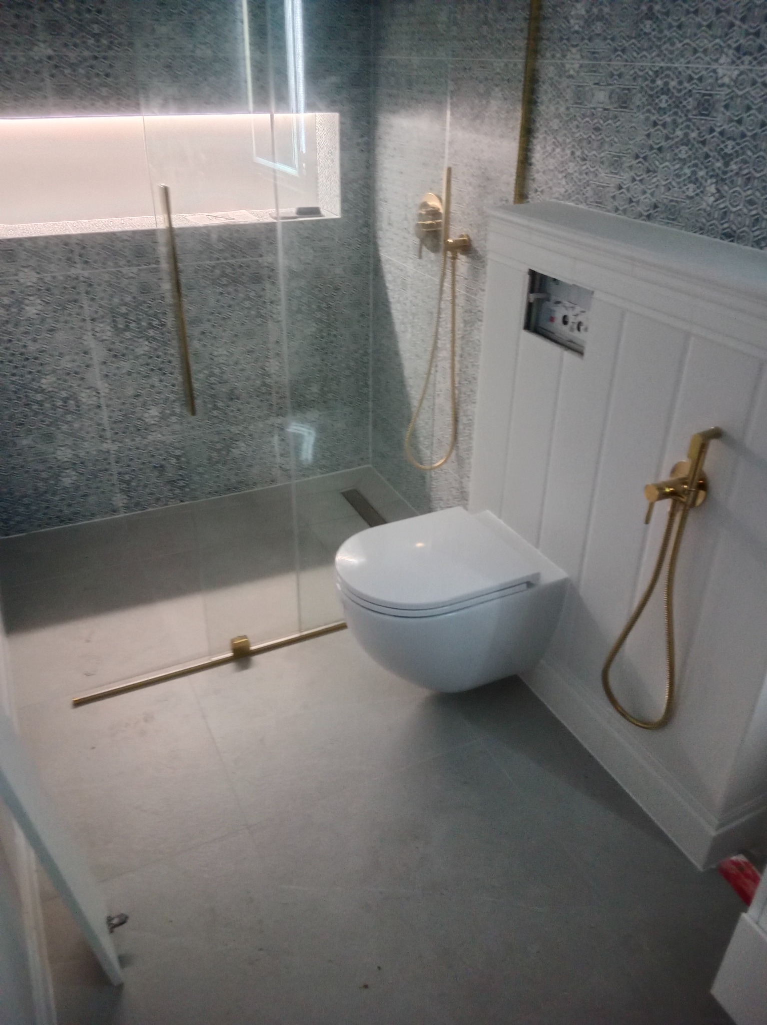 Nowoczesna toaleta podwieszana z złotą armaturą w wykończonej łazience w Warszawie. Szklana kabina prysznicowa i wzorzyste płytki dodają elegancji.