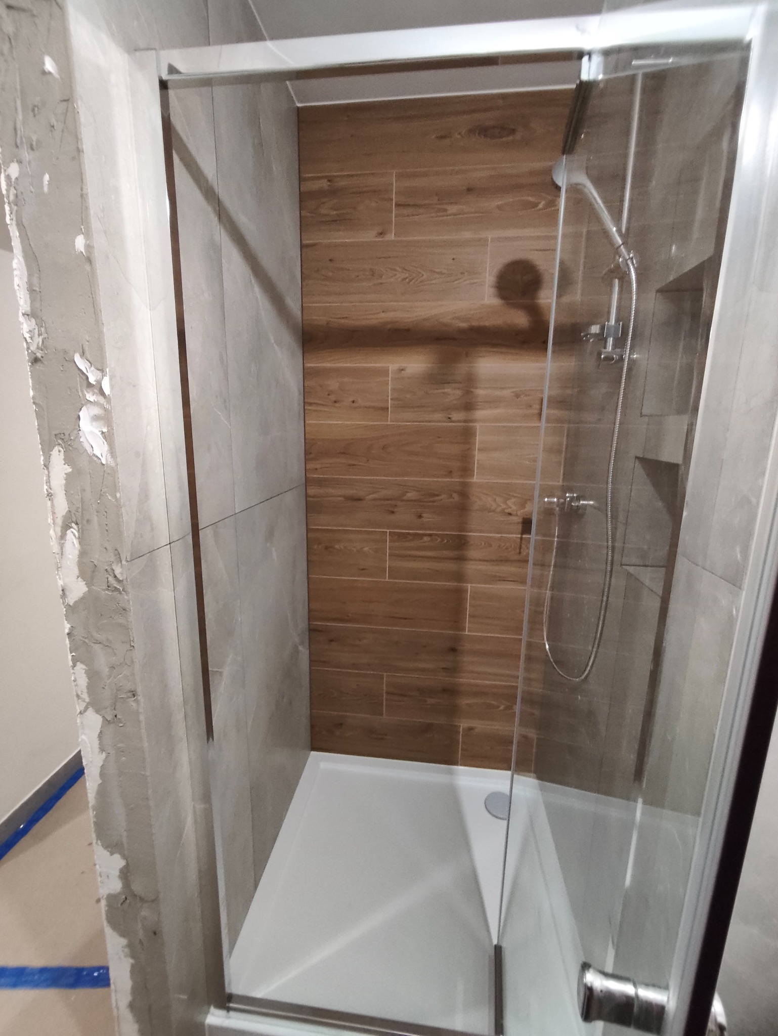 Kabina prysznicowa z drewnopodobnymi płytkami na ścianie, widoczny brodzik i szklane drzwi. Częściowo wykończona ściana z szarymi płytkami.