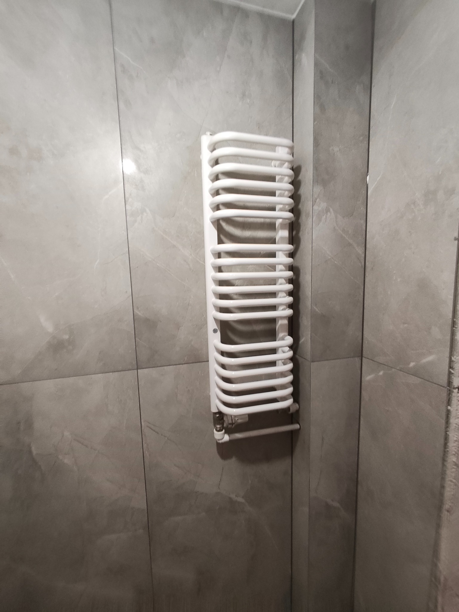Biały, drabinkowy grzejnik łazienkowy na ścianie wyłożonej szarymi, marmurkowymi płytkami. Widoczny fragment narożnika pomieszczenia.