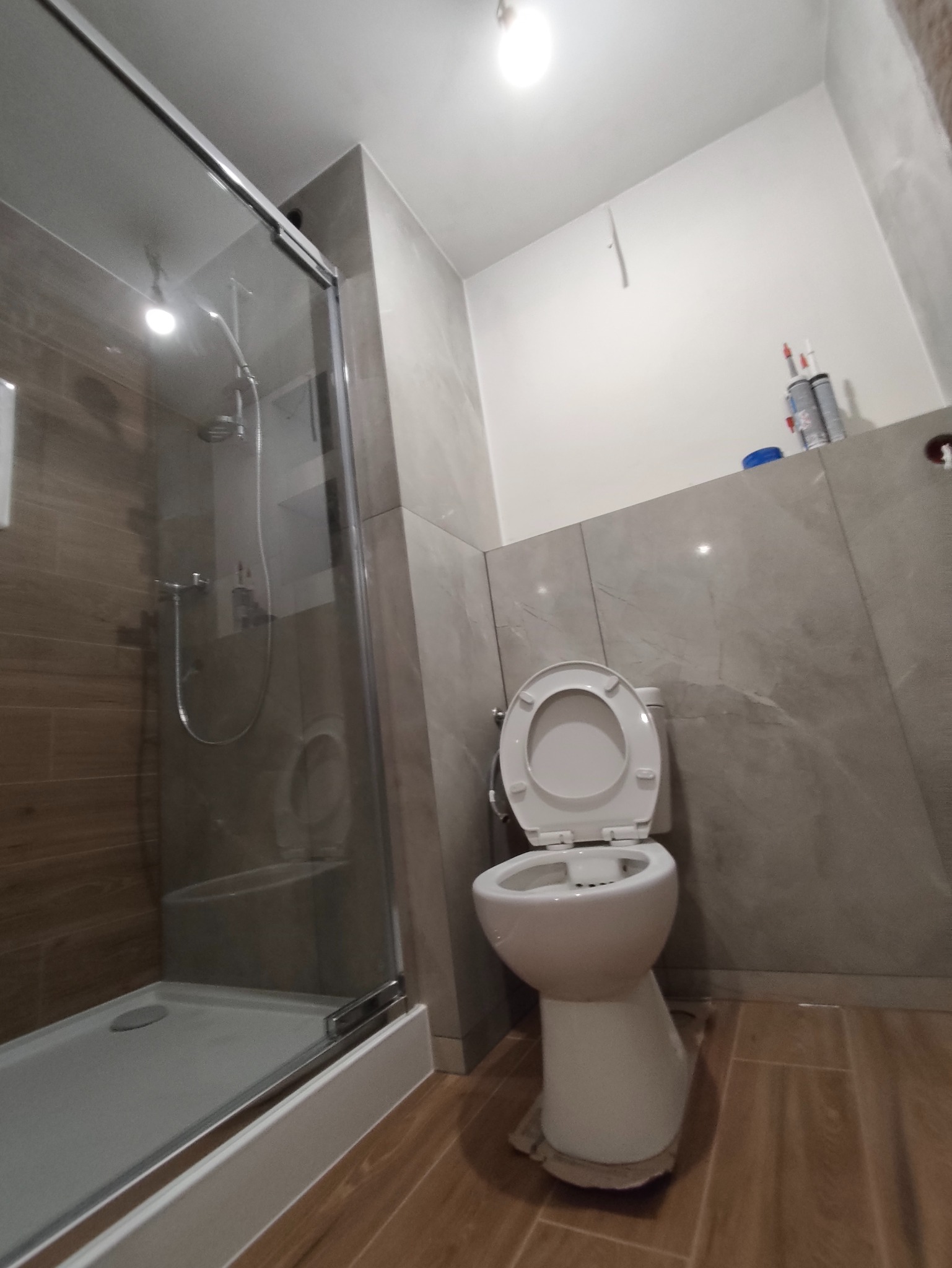 Wykończona łazienka z kabiną prysznicową i toaletą. Płytki imitujące drewno na podłodze, szare płytki na ścianach. Widok z dołu, oświetlenie górne.