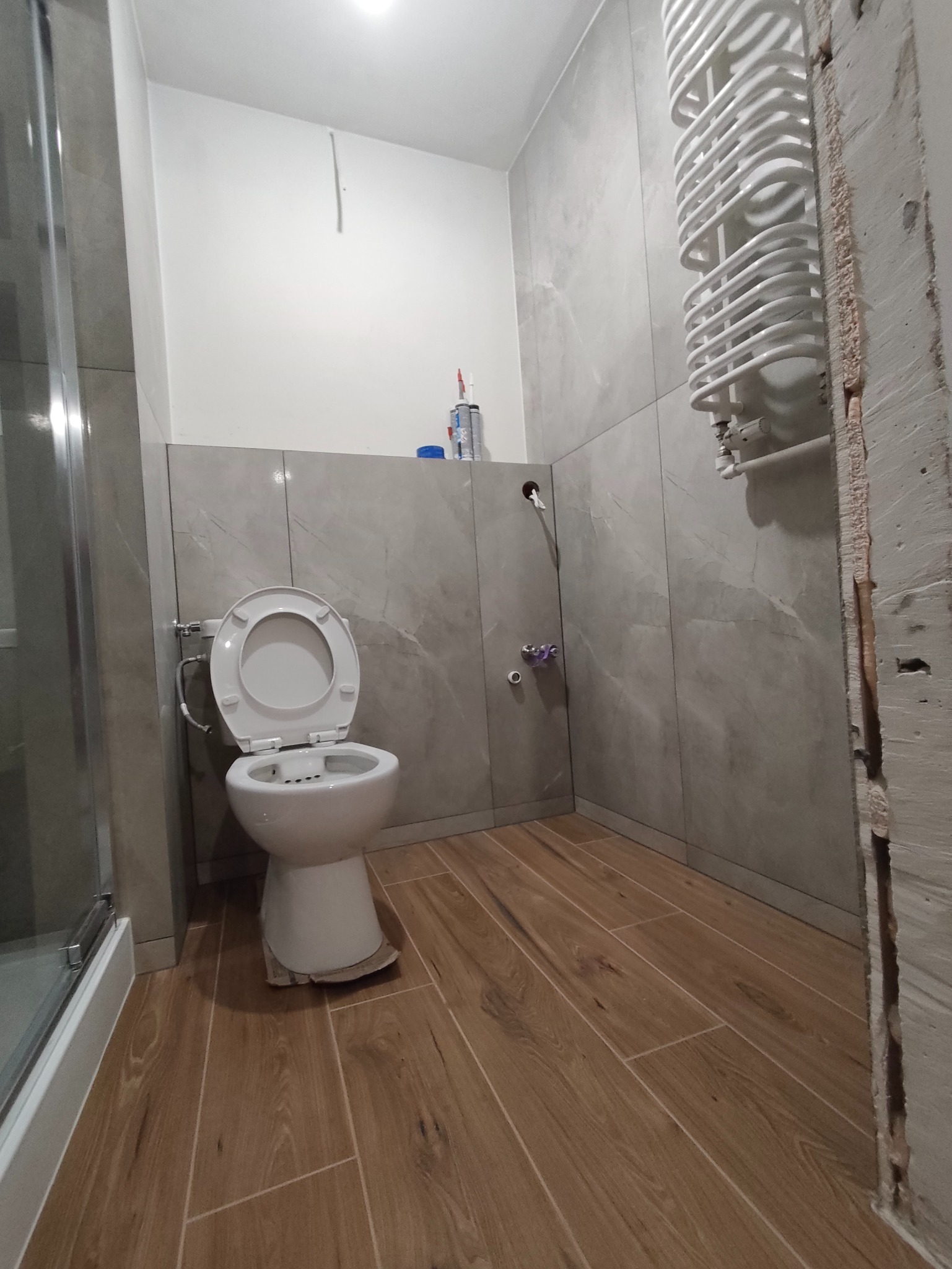 Wykończona łazienka z szarymi płytkami imitującymi marmur, drewnopodobną podłogą i białym grzejnikiem drabinkowym. Widoczny fragment kabiny prysznicowej i toaleta.