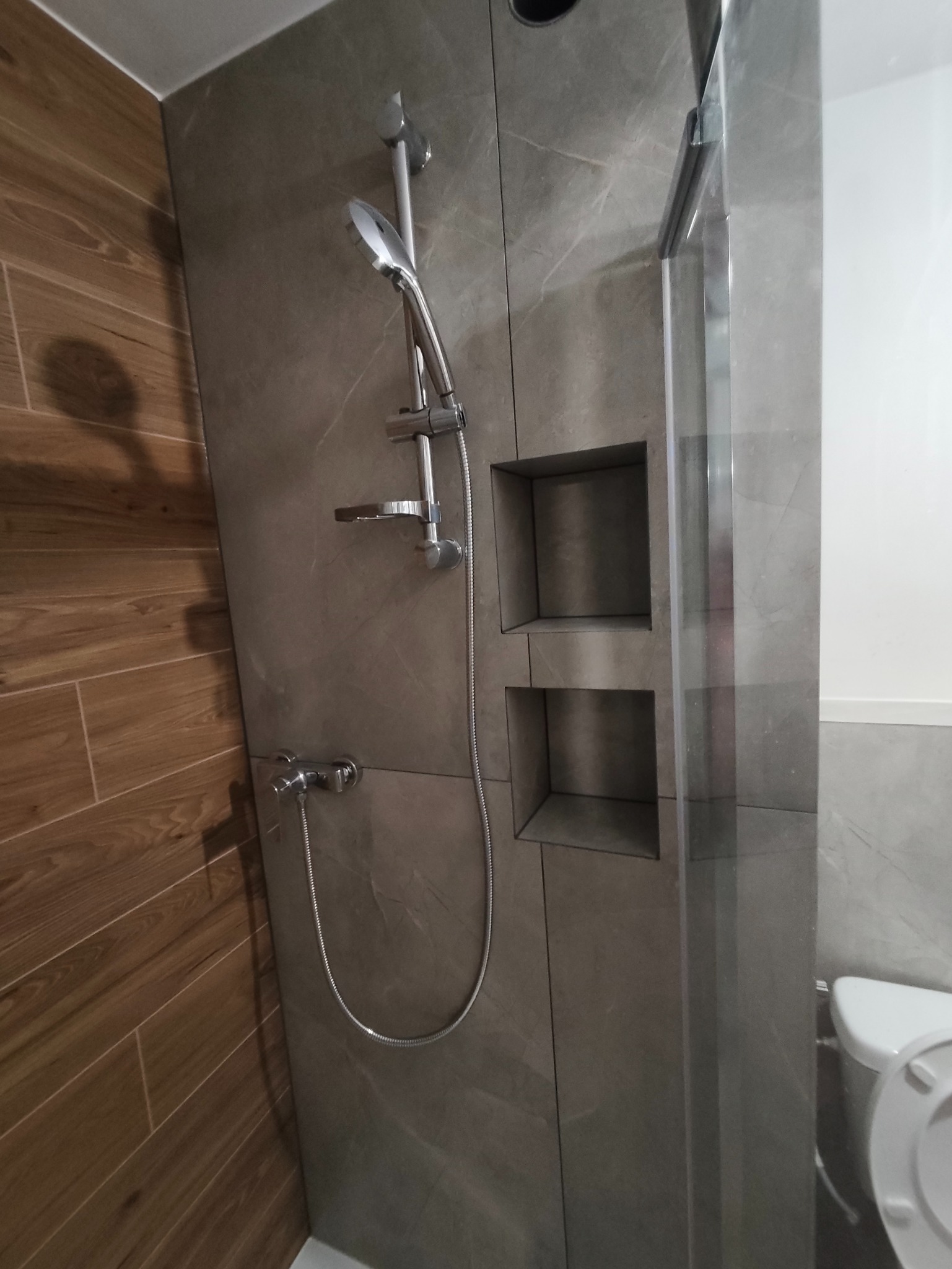 Nowoczesna kabina prysznicowa z szarymi płytkami imitującymi marmur i drewnopodobnymi płytkami na ścianie. Wnęki w ścianie prysznica. Chromowana armatura.
