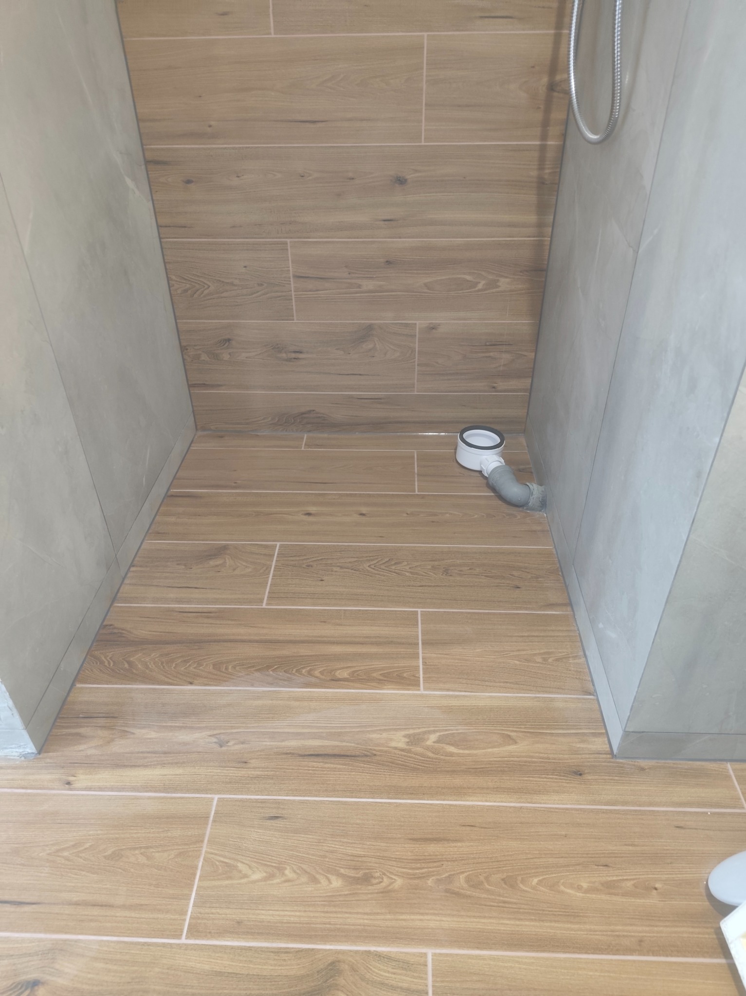 Wykończona kabina prysznicowa z drewnopodobnymi płytkami na podłodze i szarymi płytkami na ścianach. Widoczny odpływ liniowy. Minimalistyczny design.