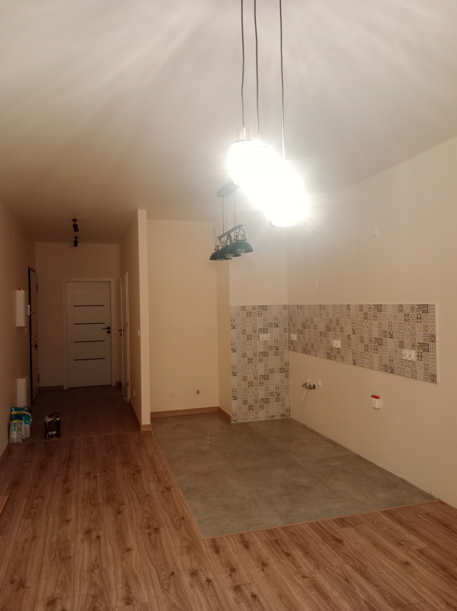 Wnętrze po częściowym remoncie: ułożona glazura na ścianie i podłodze w kuchni, widoczne przejście do parkietu w salonie, lampa wisząca.