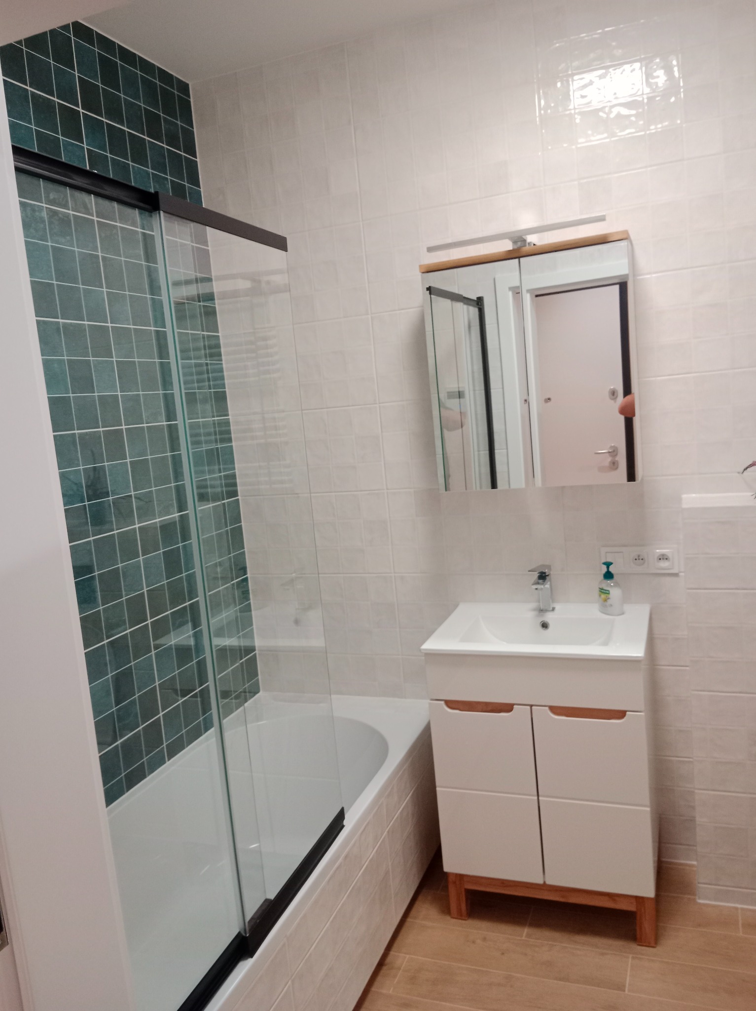 Nowoczesna łazienka z kabiną prysznicową z zielonymi płytkami, białą umywalką z szafką i lustrem z oświetleniem. Jasne płytki na ścianach i drewnopodobna podłoga.