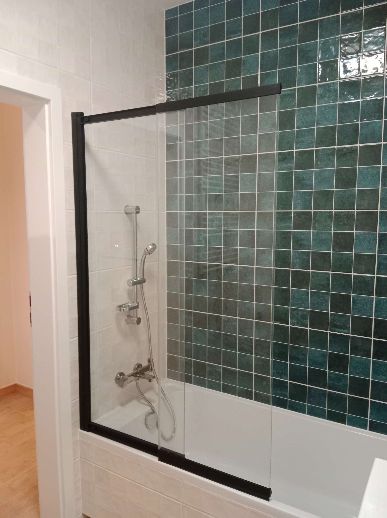 Wykończona łazienka z kabiną nawannową w czarnej ramie i zielono-białymi płytkami. Widoczna armatura i fragment podłogi. Nowoczesny design.