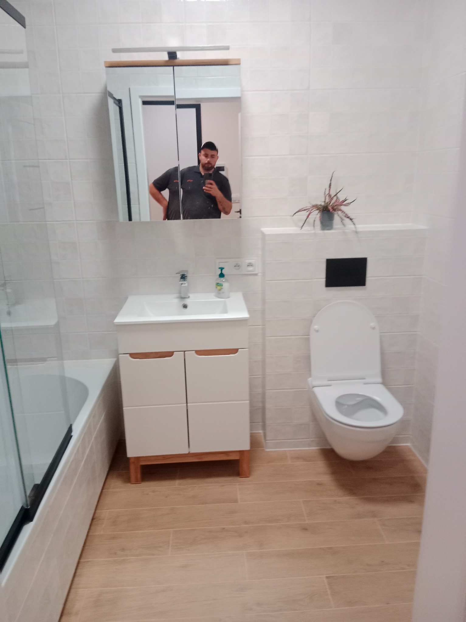 Jasna łazienka po remoncie: białe płytki, drewnopodobna podłoga, biała szafka z umywalką i toaleta. W lustrze odbija się osoba robiąca zdjęcie.