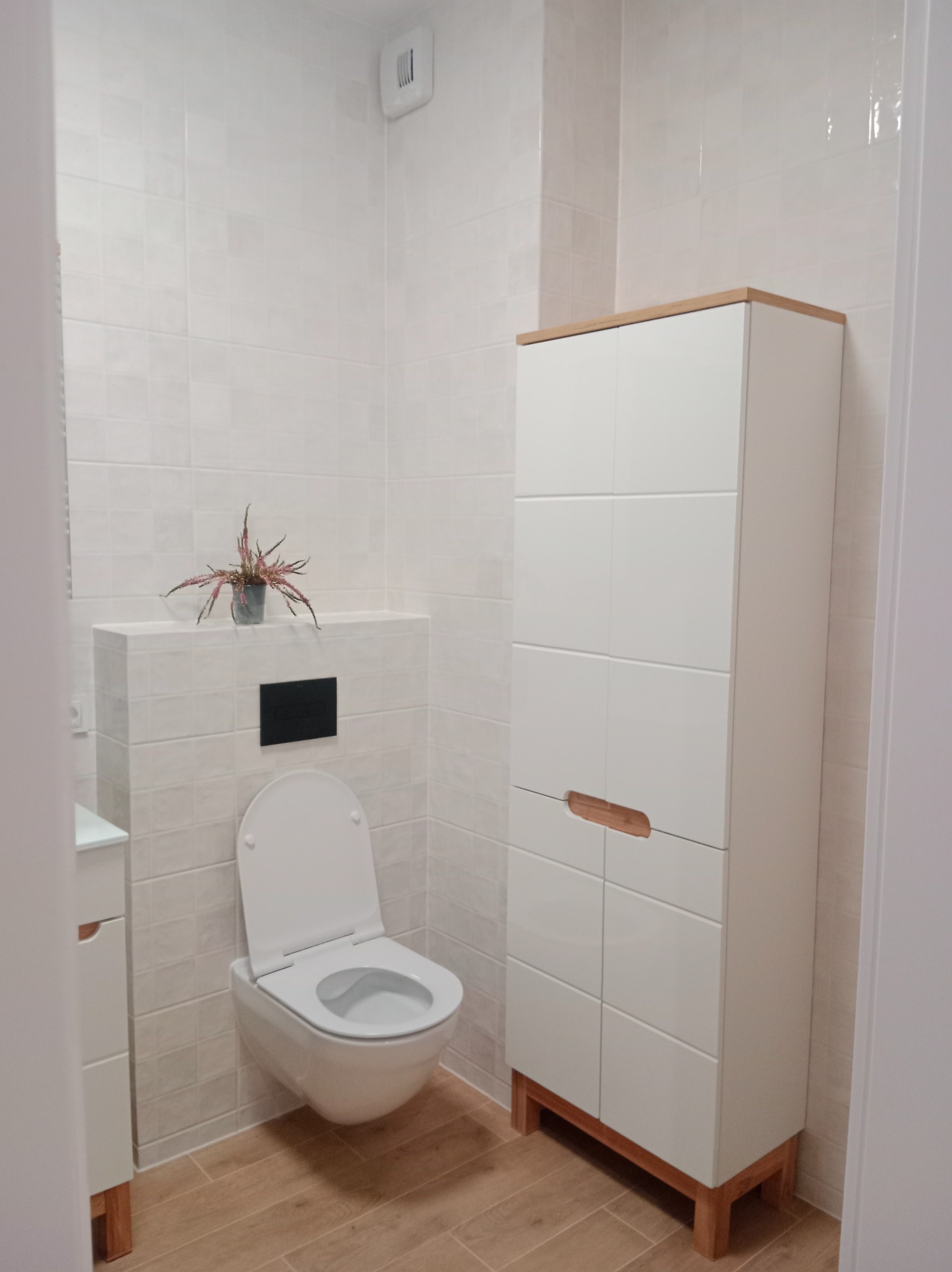 Jasna łazienka po remoncie: białe płytki, podwieszana toaleta, szafka z drewnianymi akcentami i drewnopodobna podłoga. Minimalistyczny styl.