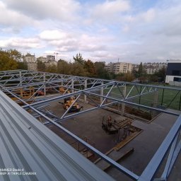 KS DACH SYSTEM DARIA ŚLUBOWSKA - Konstrukcja stalowa dachu w trakcie montażu, widok z góry na plac budowy z maszynami i elementami konstrukcyjnymi na tle zabudowy miejskiej i zieleni.