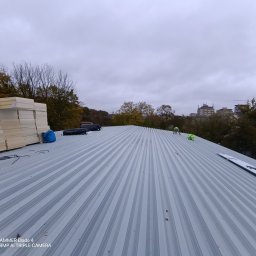 KS DACH SYSTEM DARIA ŚLUBOWSKA - Montaż blachy trapezowej na dachu z widocznym ociepleniem. Prace dekarskie w toku, widok z góry na szary dach i pracowników w oddali.