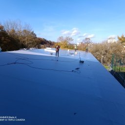 KS DACH SYSTEM DARIA ŚLUBOWSKA - Ekipa w pracy na płaskim dachu pokrytym szarą membraną, widoczne narzędzia i materiały izolacyjne. W tle drzewa i budynki w oddali.