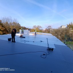 KS DACH SYSTEM DARIA ŚLUBOWSKA - Prace na dachu: ekipa w kaskach montuje izolację termiczną na płaskim dachu budynku, widoczne narzędzia i materiały izolacyjne. W tle drzewa i budynki.